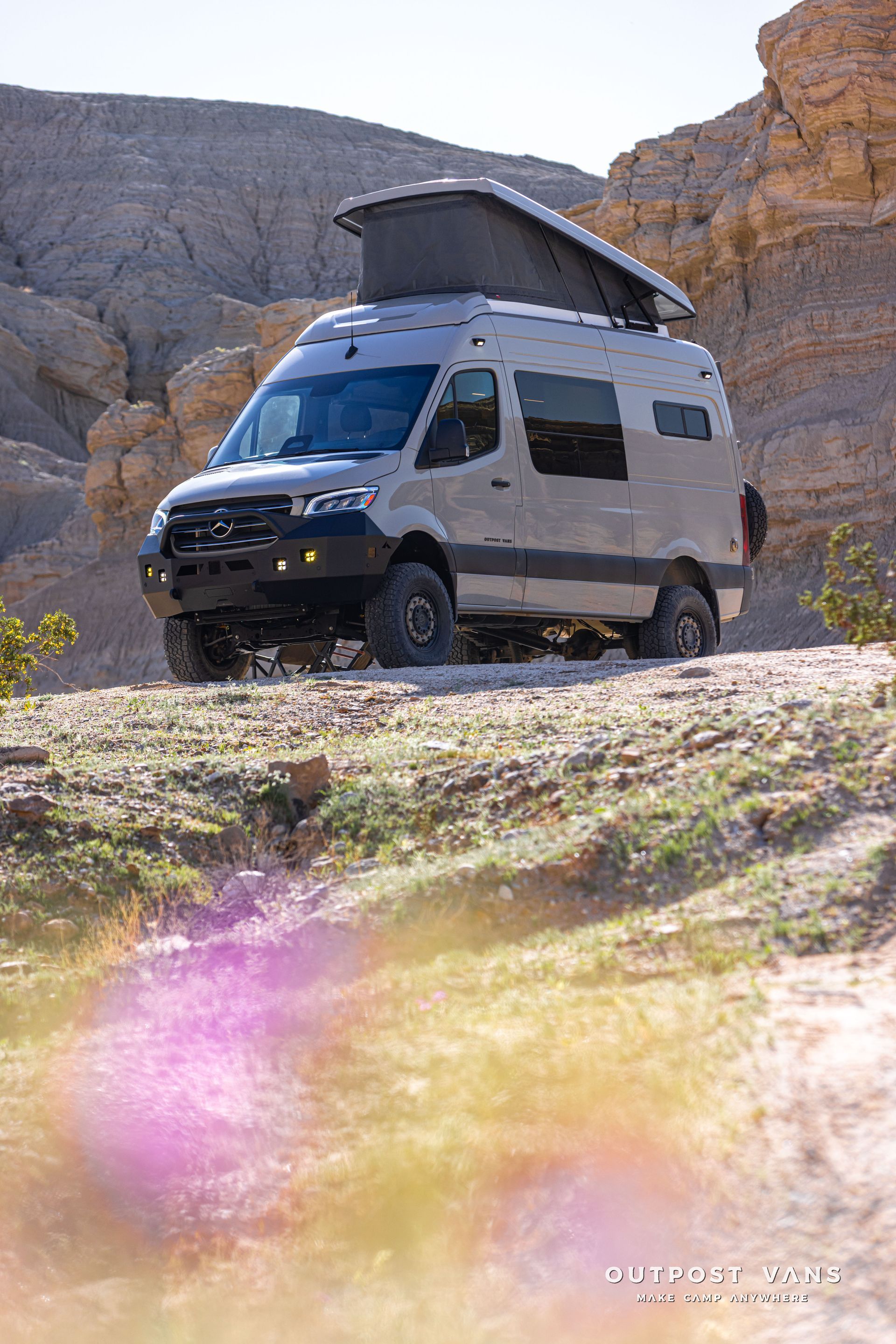 White camper van with pop-up roof parked on rocky desert terrain beside cliffs.