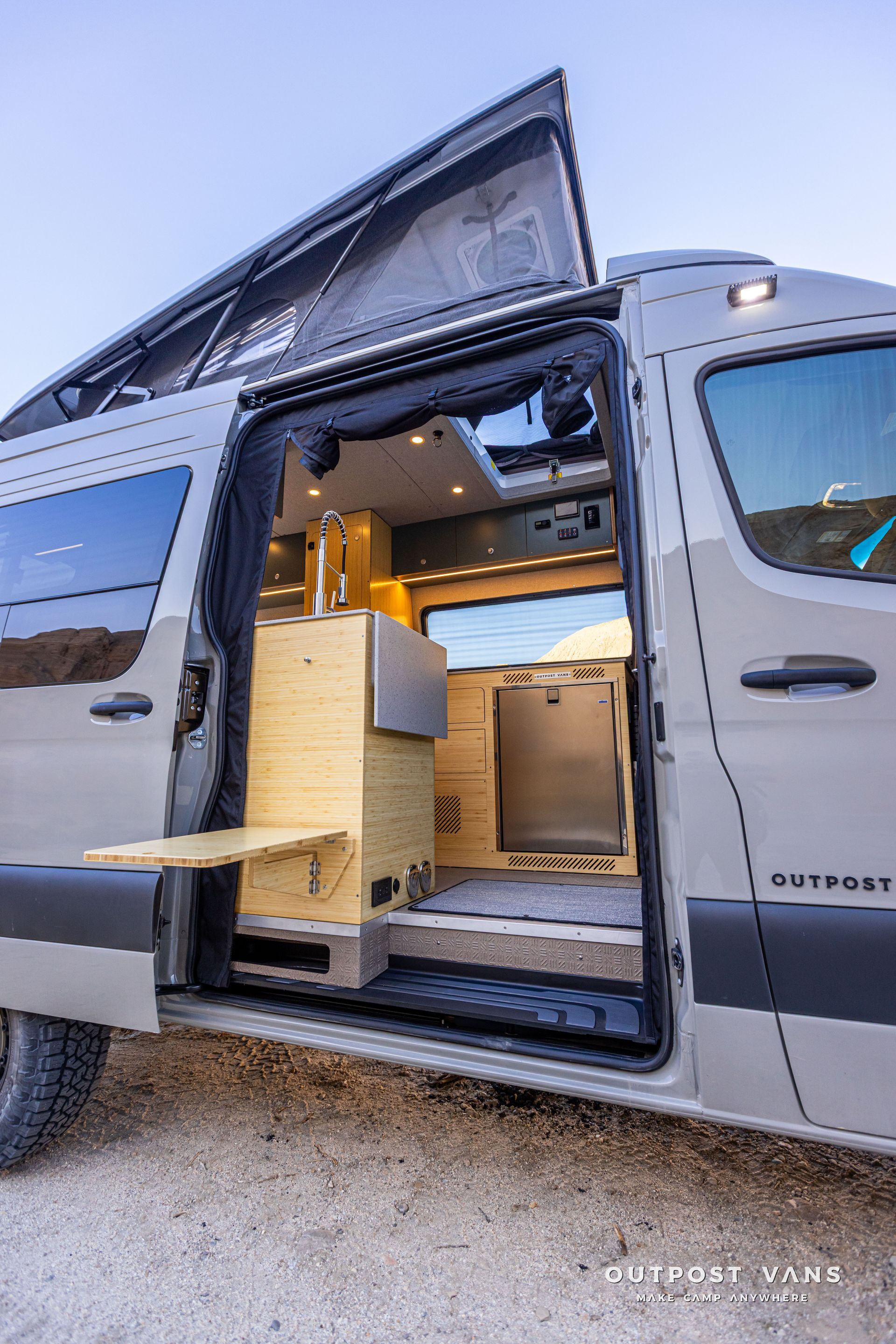 Gray camper van with pop-up roof and open side door revealing a wooden interior on a gravel lot