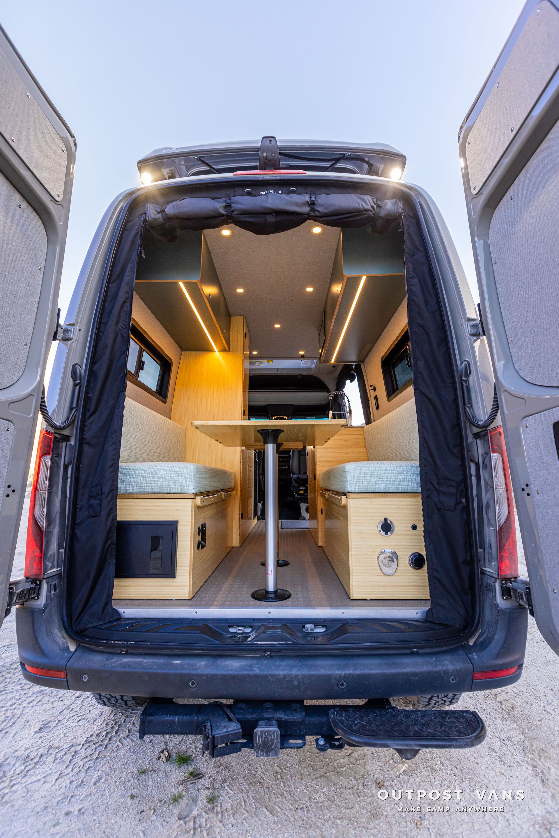 Open van with custom camper interior, warm wood cabinets, benches, and central table viewed from the rear.