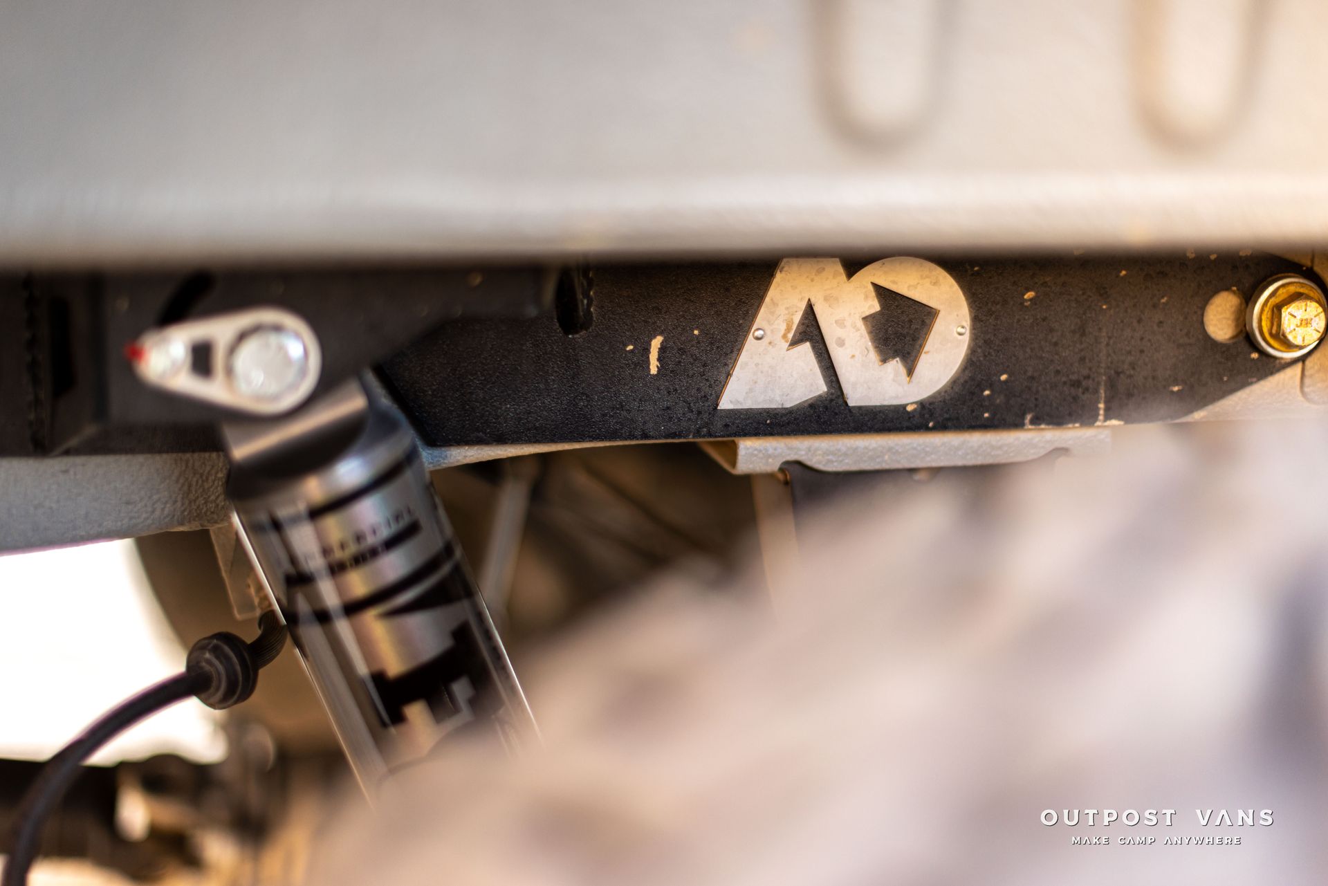 Close-up of a black suspension bracket with a gold logo and bolt, under a vehicle chassis