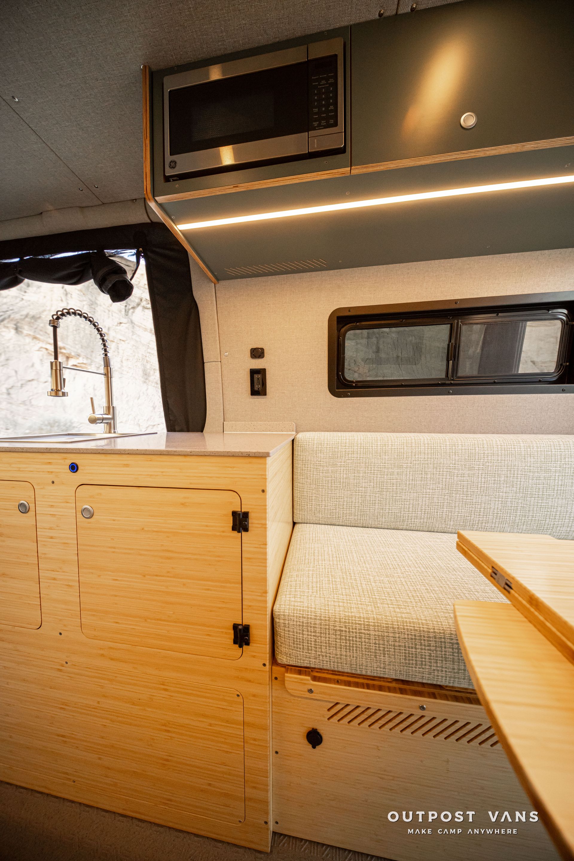 Camper van interior with wood cabinets, small sink, bench seat, window, and LED strip lighting