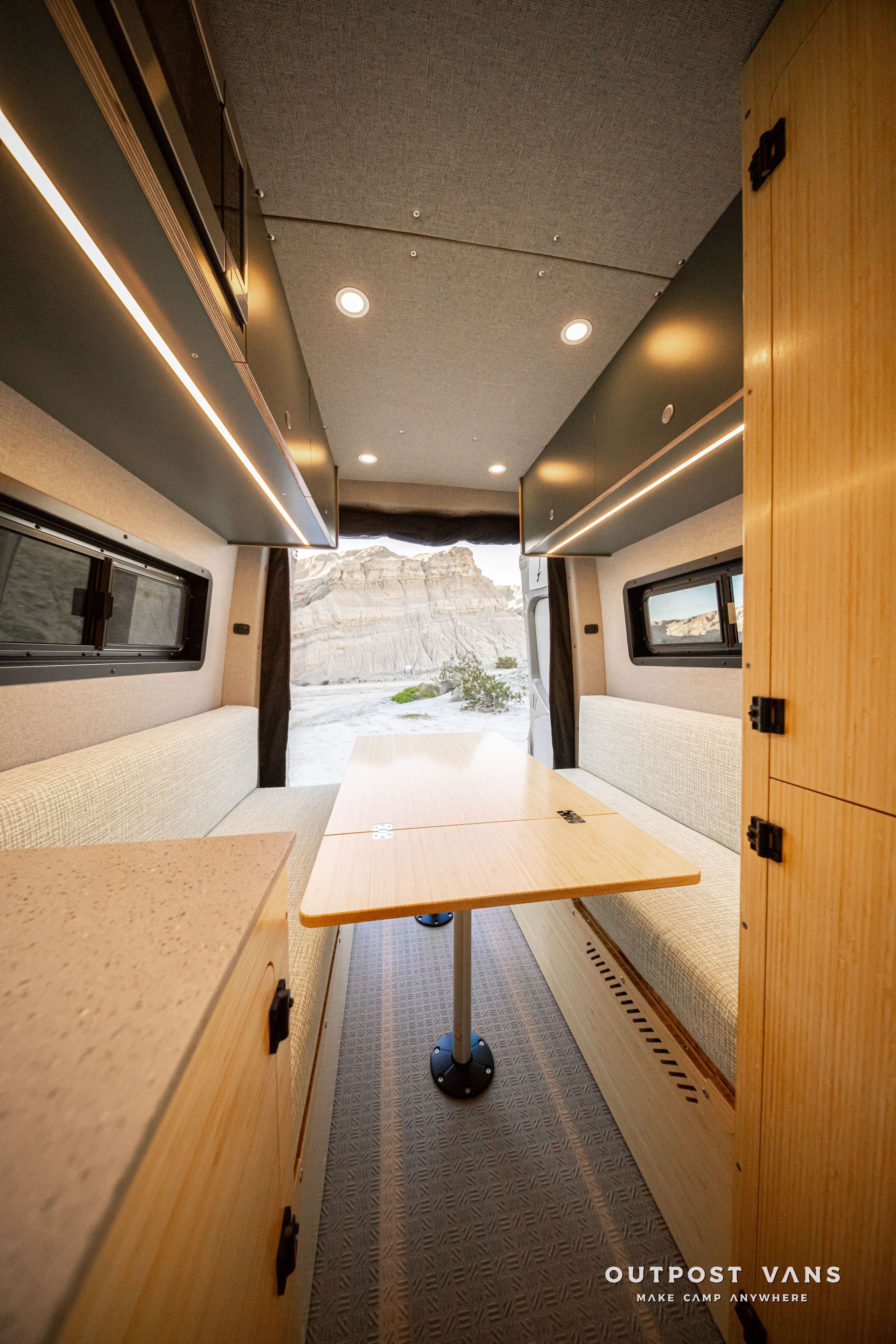 Compact camper van interior with fold-out table, wood cabinets, and open rear view to snowy mountains