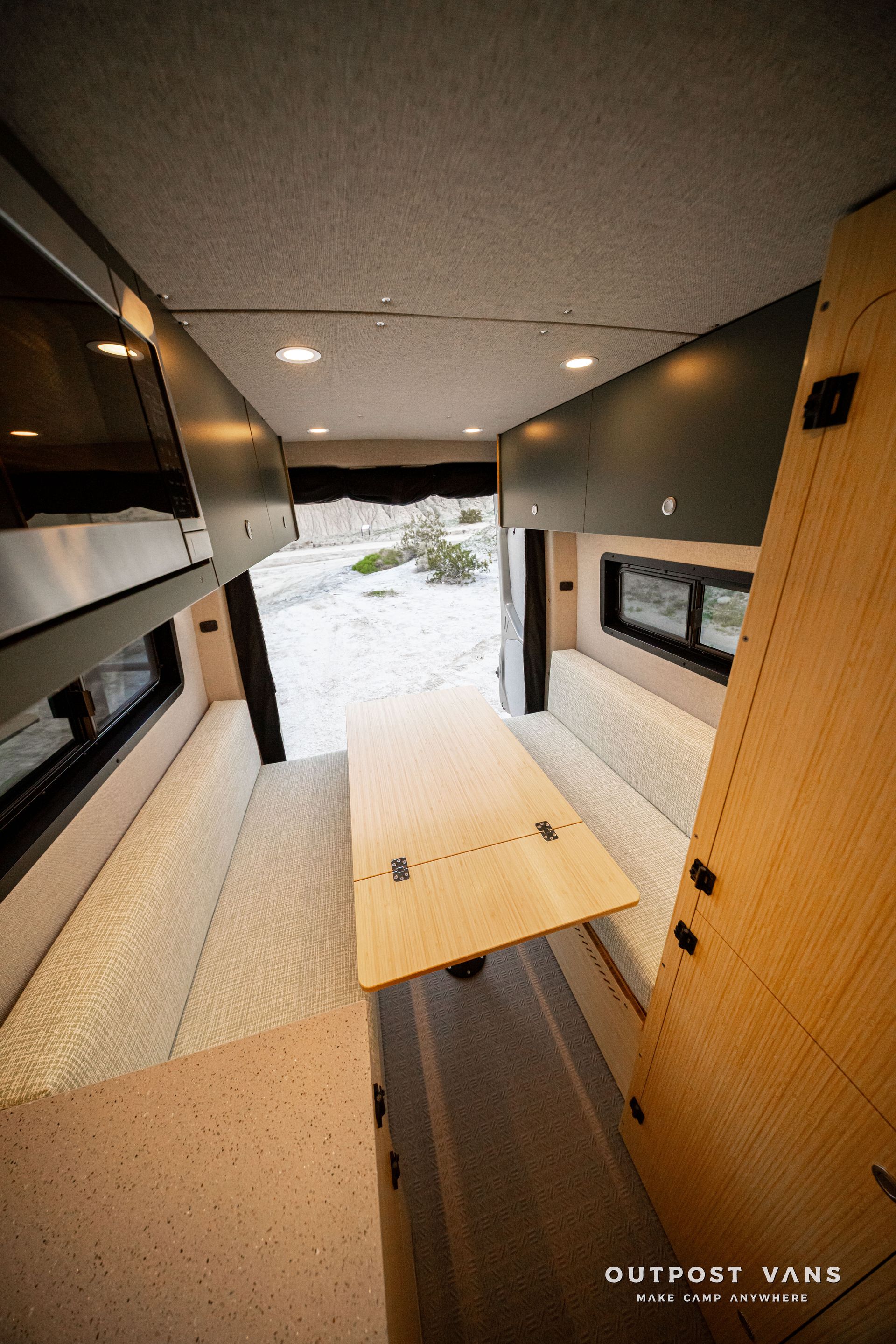 Interior of a compact camper van with wood table, bench seating, and open rear door to snowy outdoors