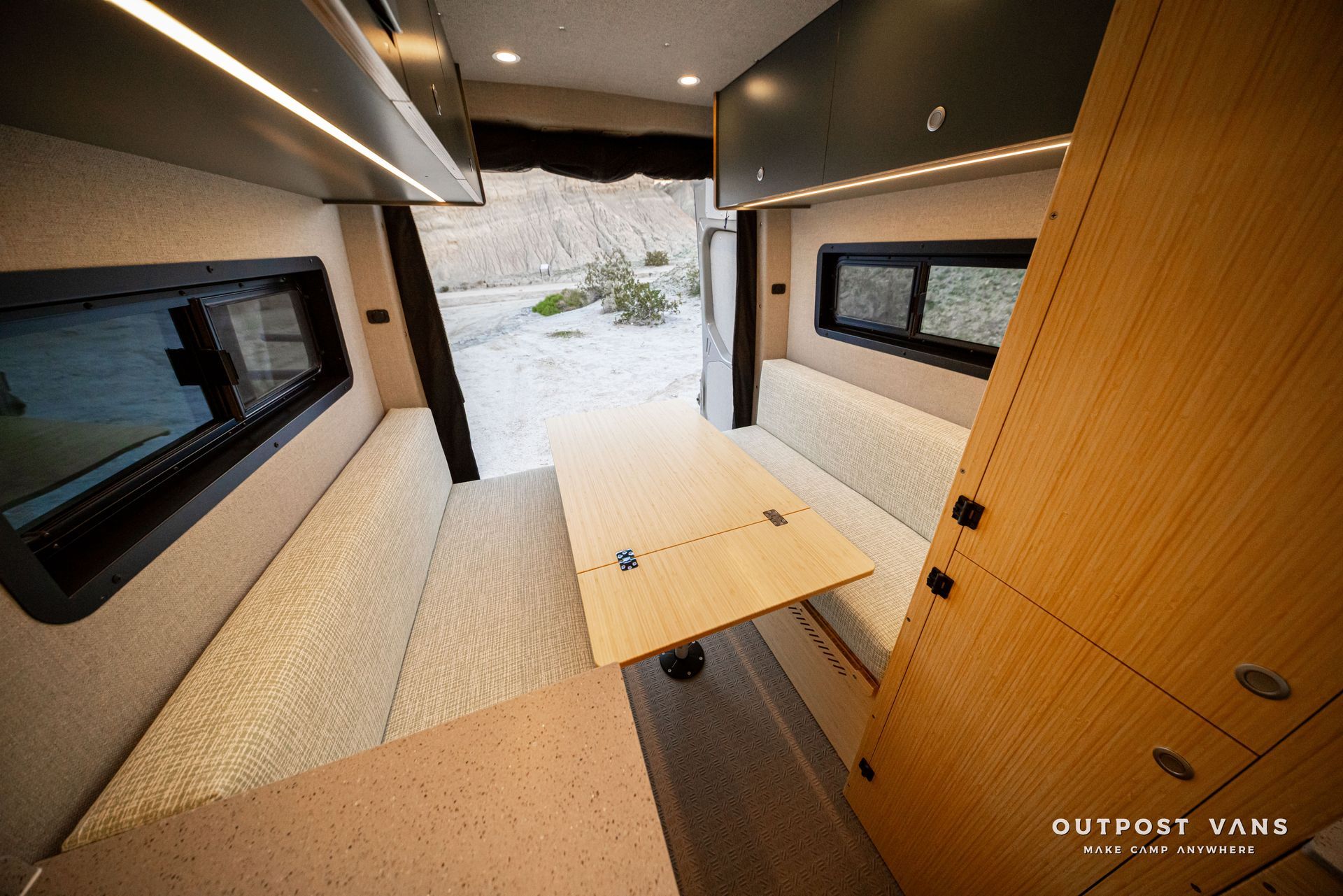 Compact camper van interior with bench seating, wood table, and open rear doors to snowy outdoors