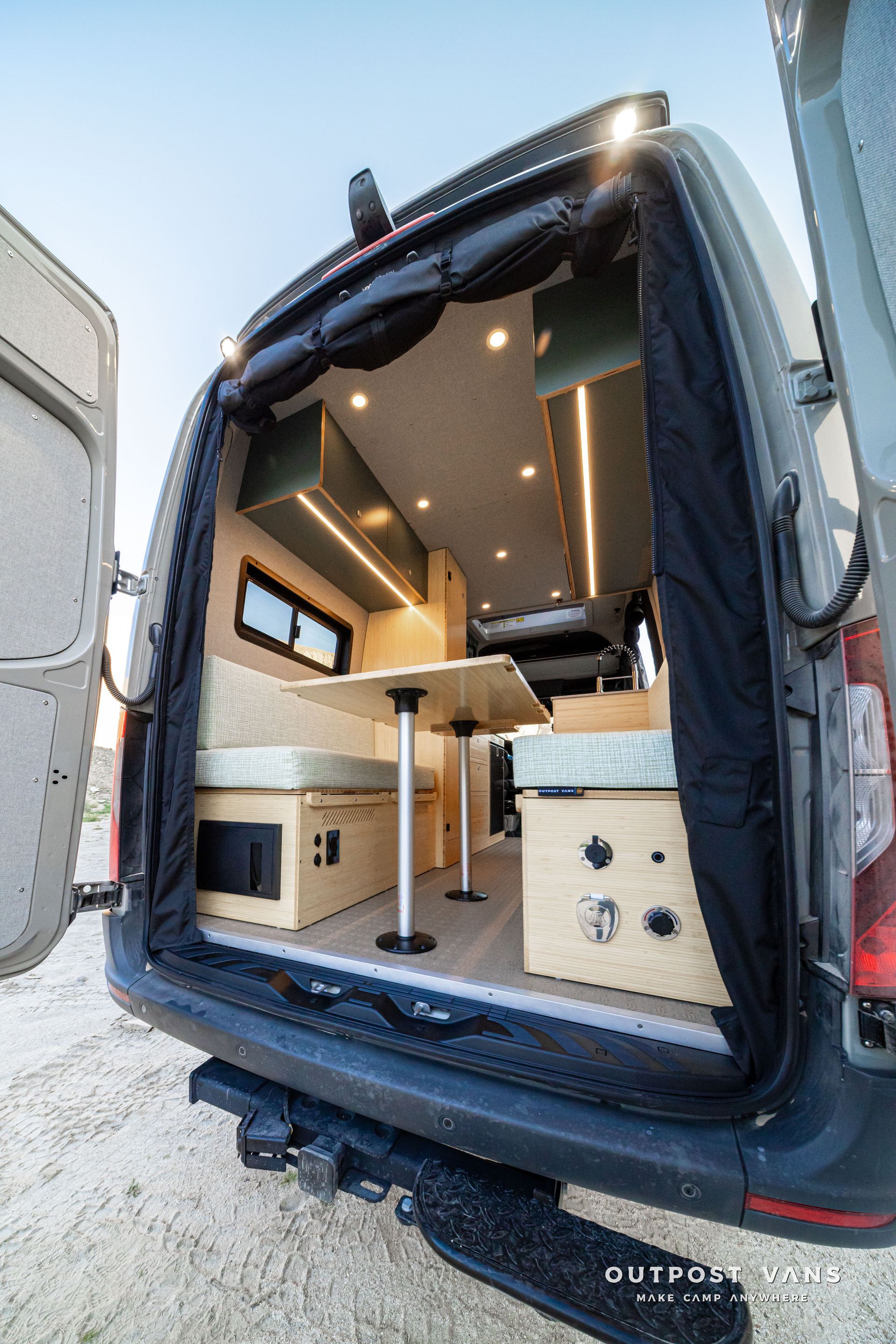 Open camper van interior with wood cabinets, a fold-out table, and bed platform at the rear door.