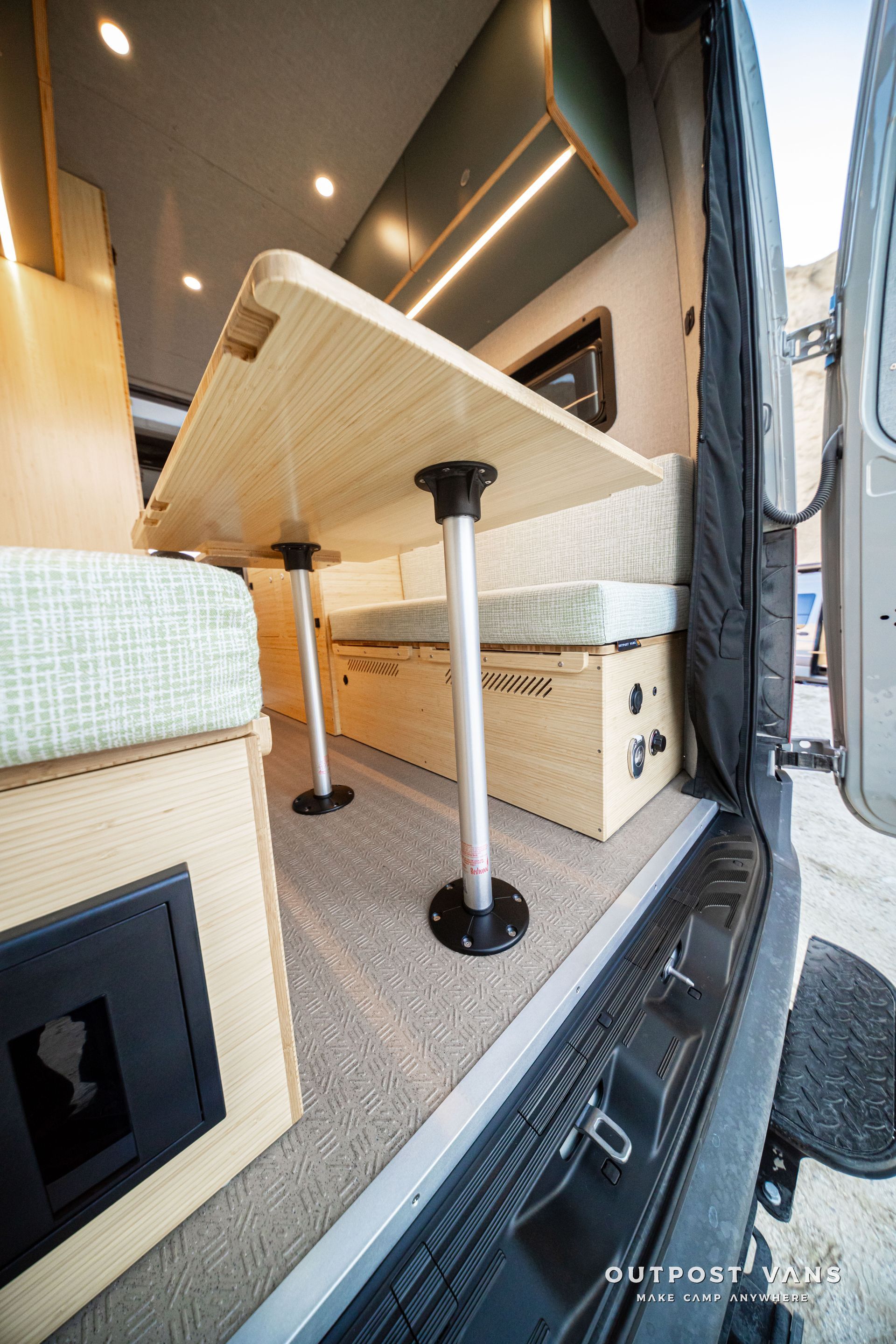Compact RV dinette with a raised tabletop, black pedestal legs, beige cushions, and a narrow walkway by the door