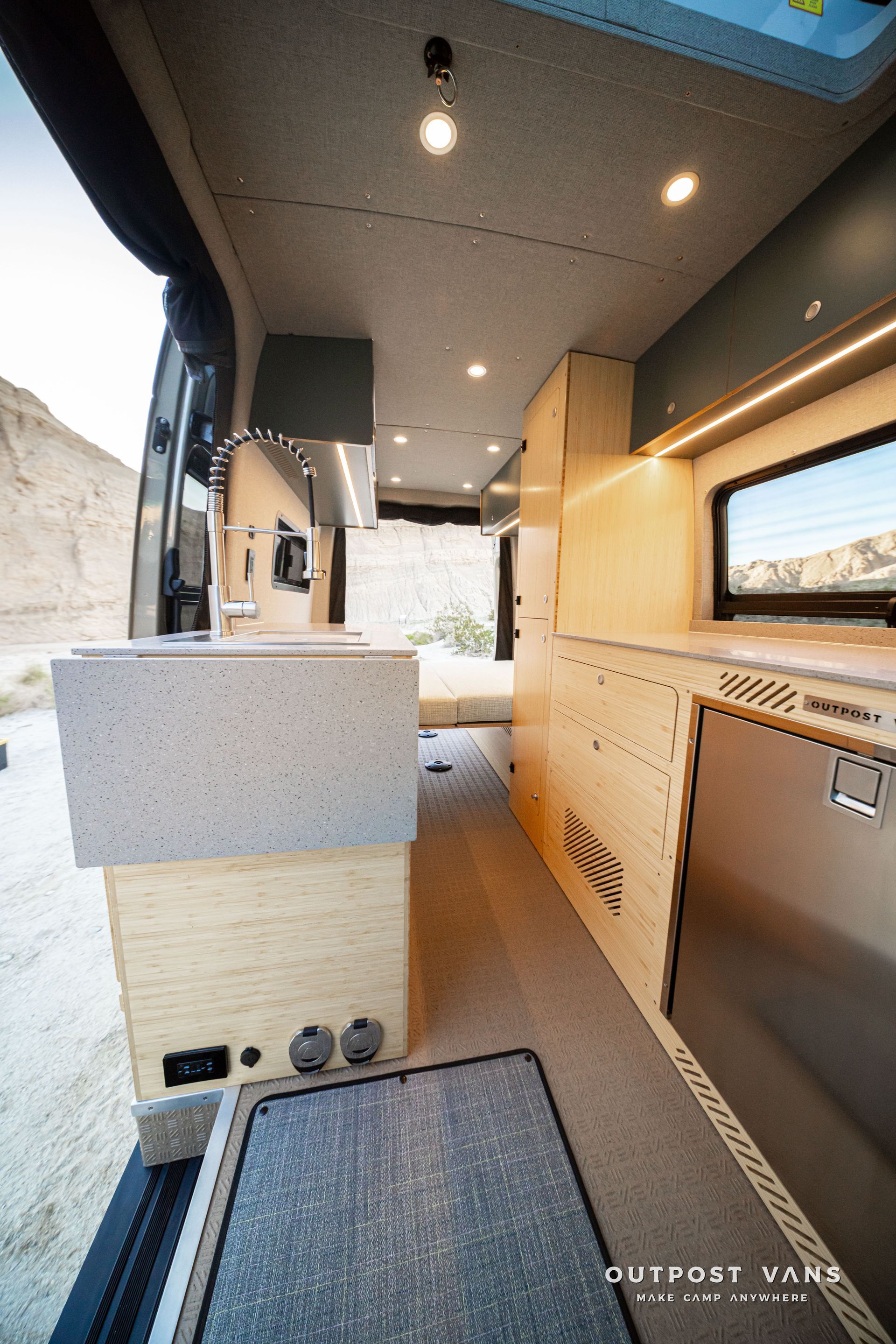 Compact outdoor kitchen in a camper van with wood cabinetry, sink, and countertop under a lit canopy.