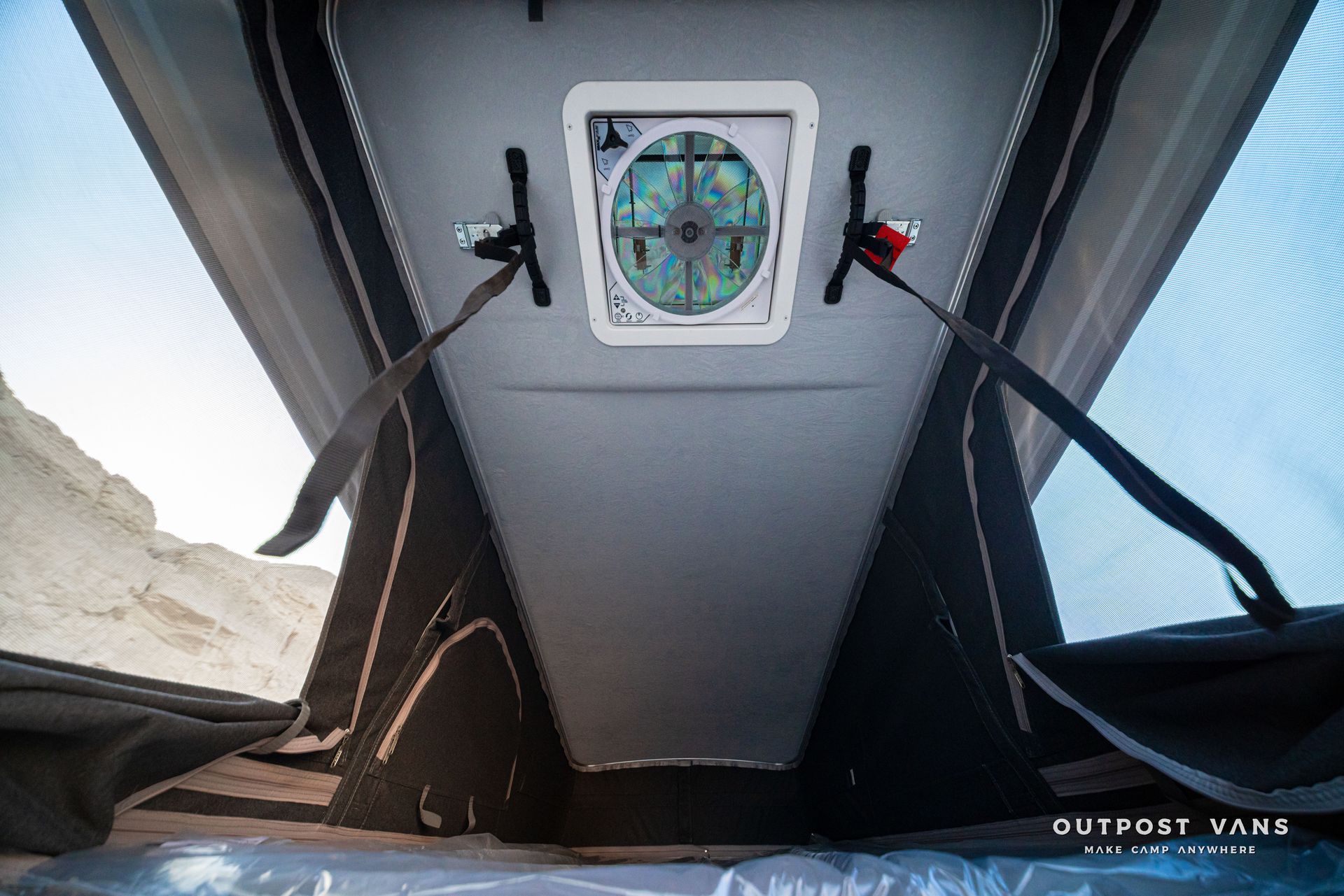 Inside a tent, a square fan vent is centered on the ceiling with straps and open mesh sides.