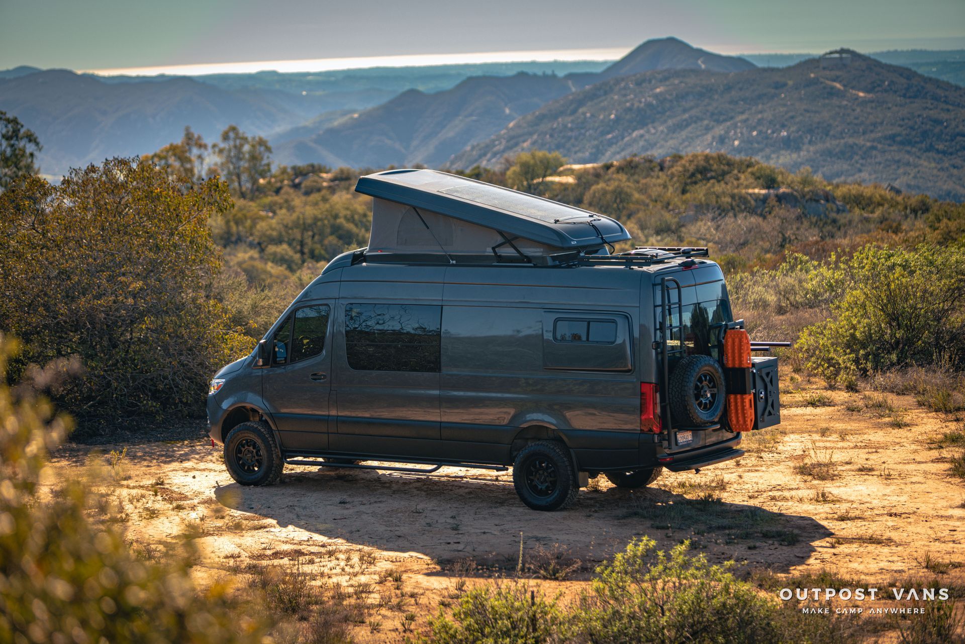 A van is parked on top of a dirt hill with mountains in the background.