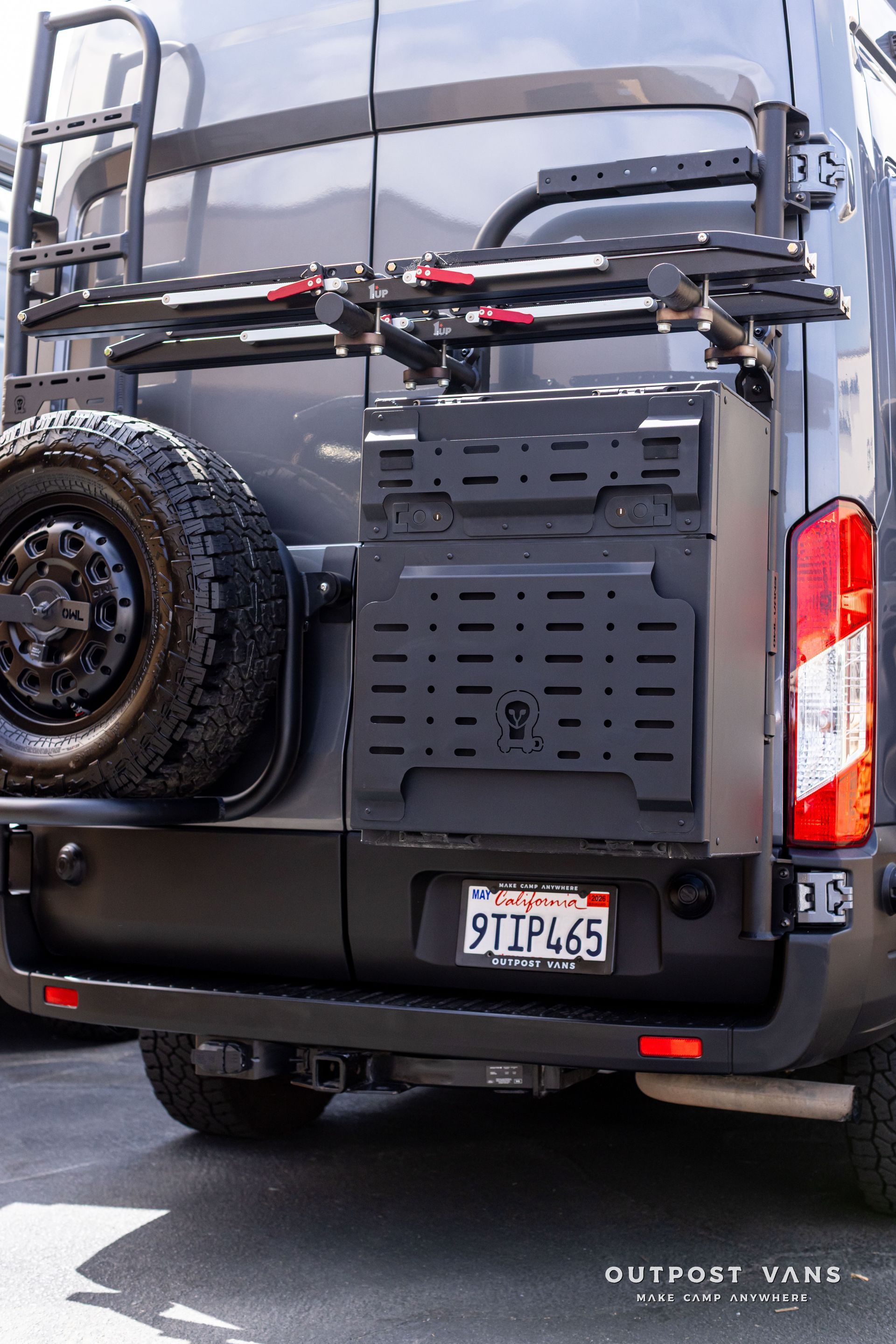 Black van rear with spare tire, cargo rack, and California plate outdoors on a dirt road