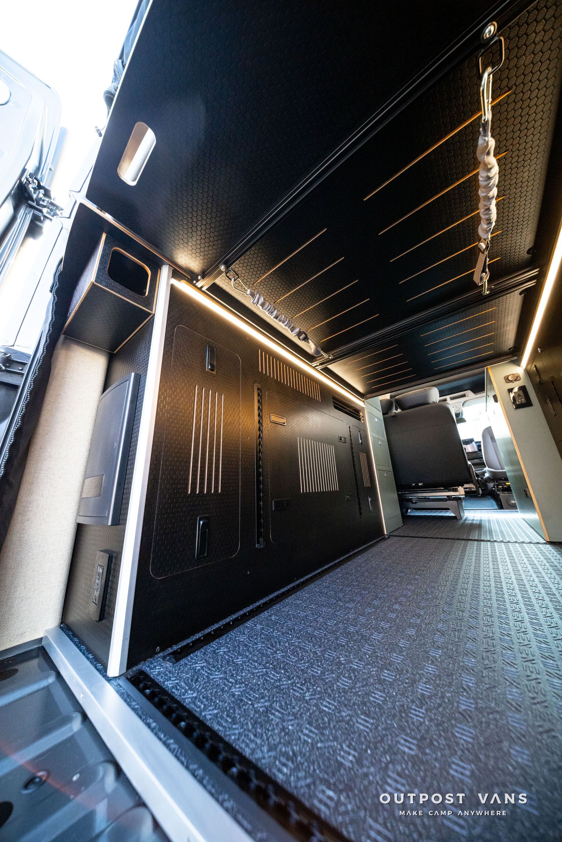 Black industrial hallway with textured floor, overhead strip lights, and an open doorway at the end.