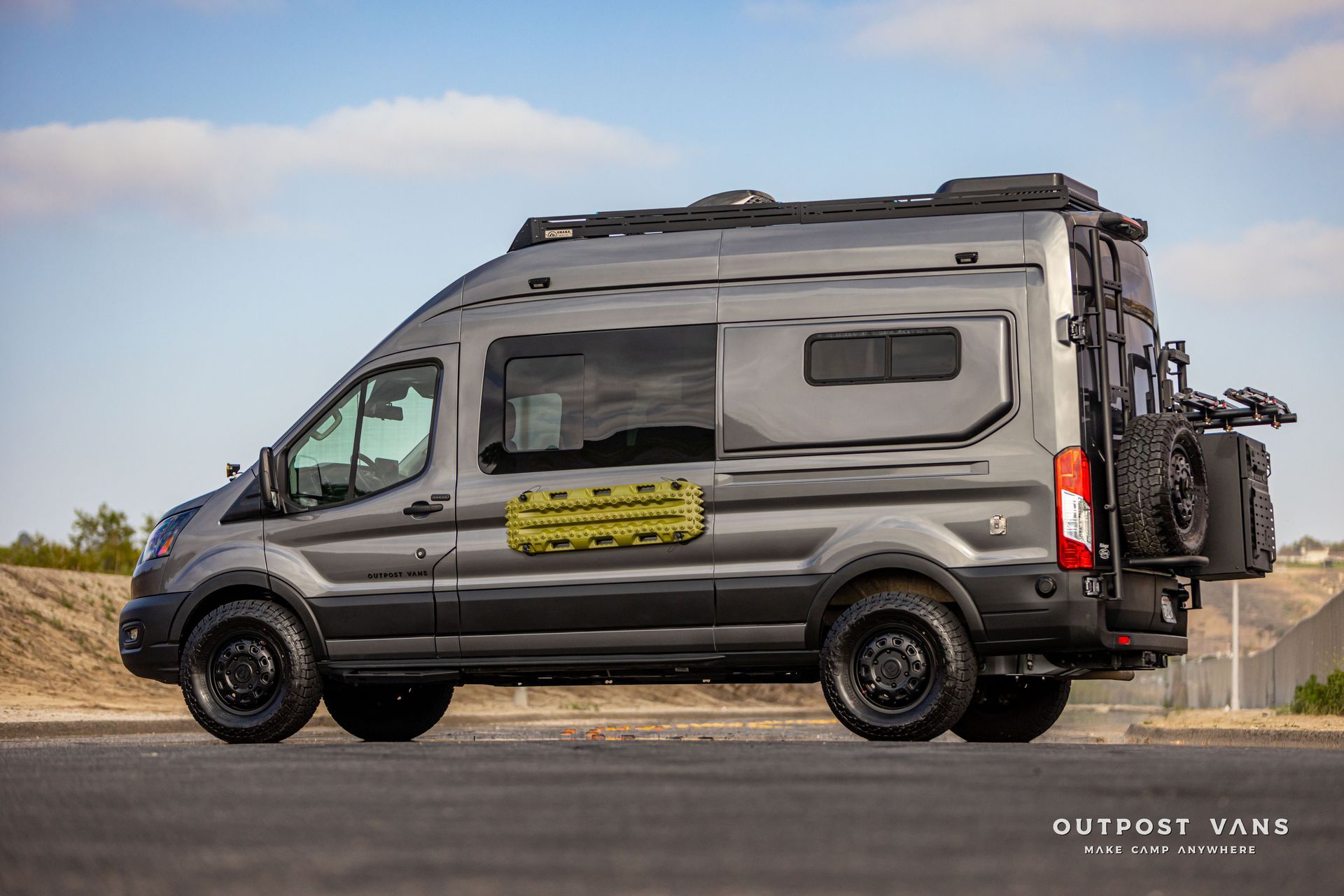 Gray camper van parked on a road, with roof gear and rear bike rack, under a clear sky.