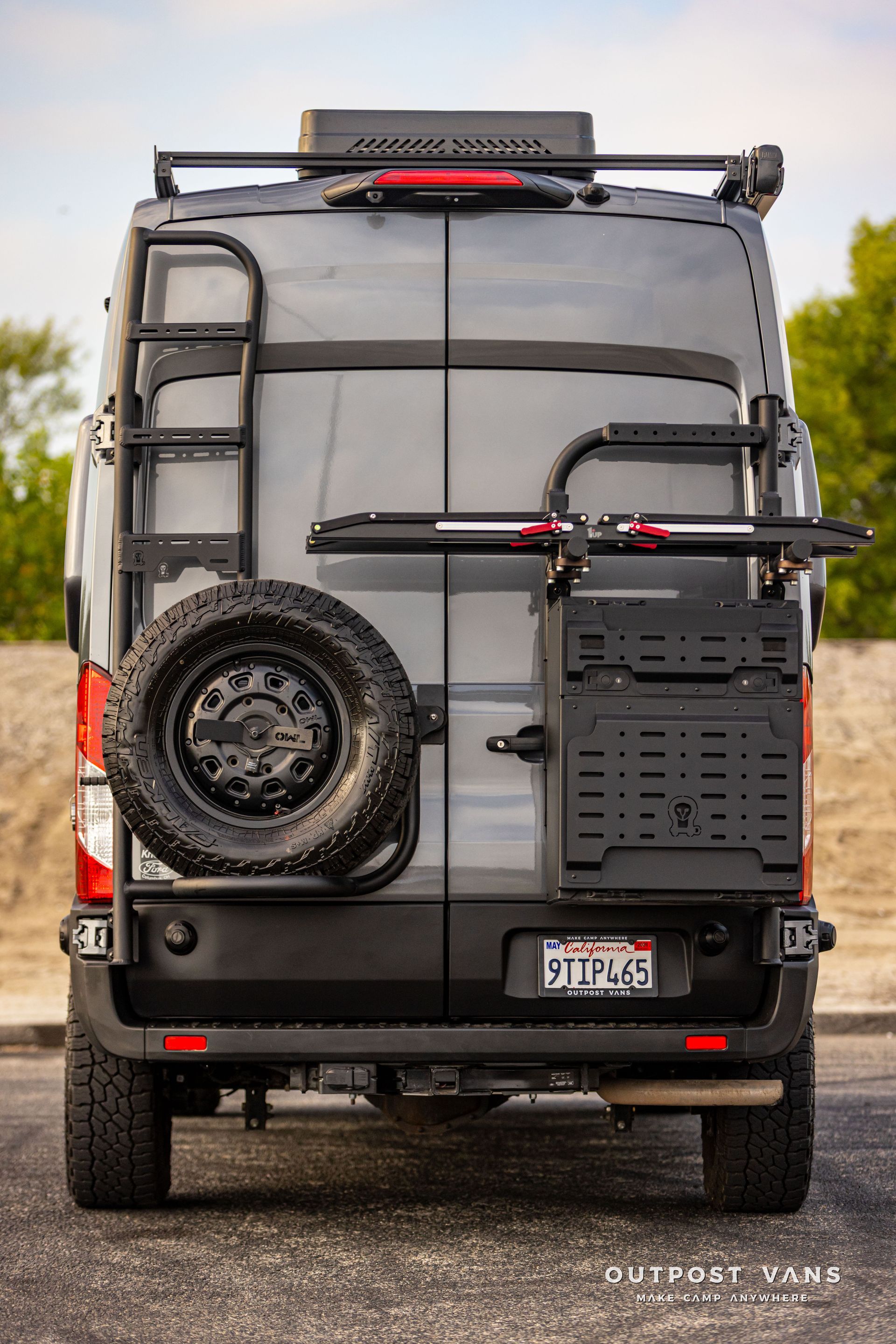 Rear view of a gray camper van with a roof rack, spare tire, and outdoor gear mounted on the back.