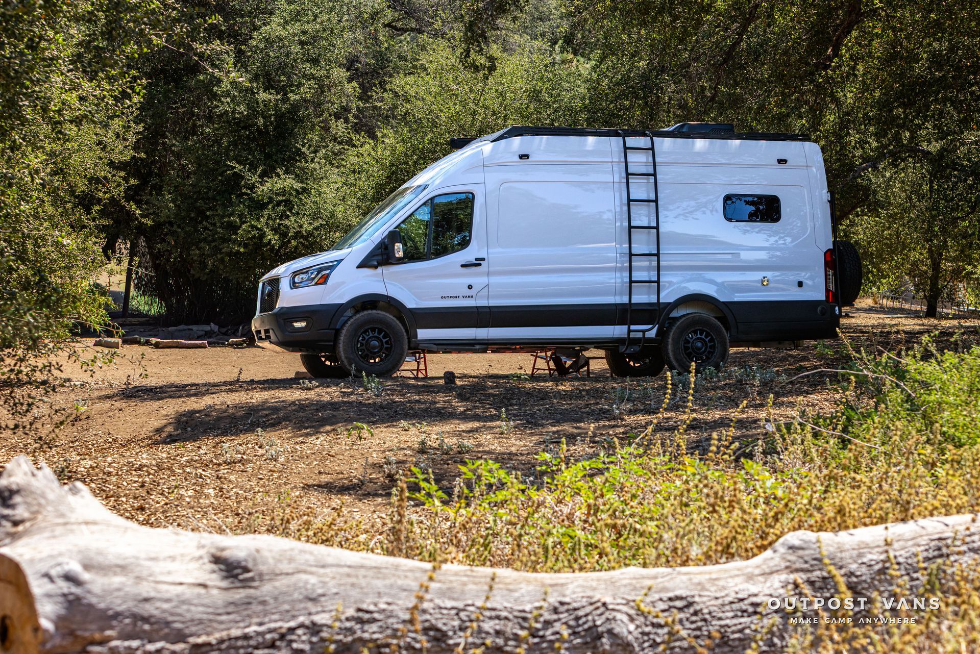 White cargo van parked on a dirt clearing beside trees, with a fallen log in the foreground.