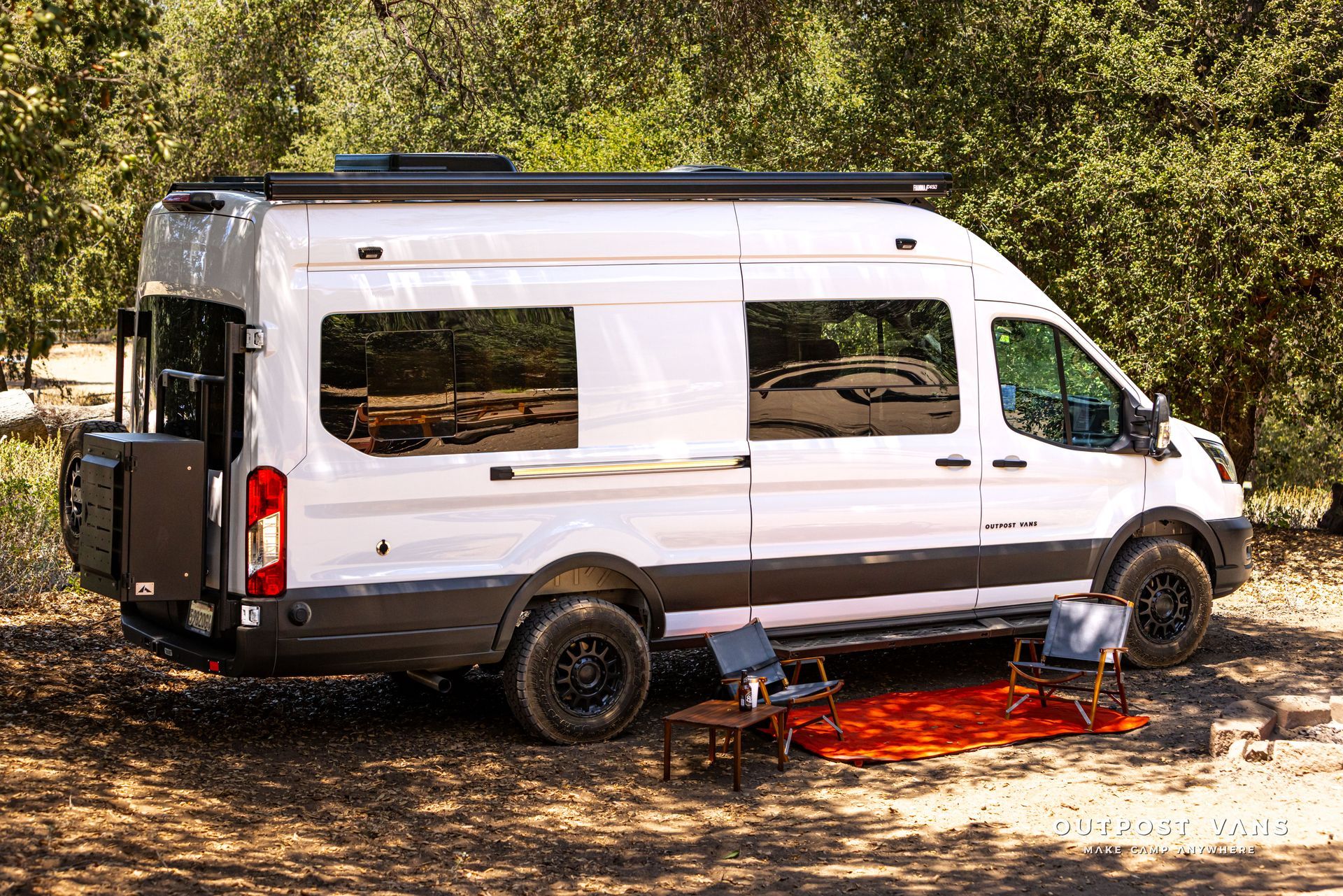 White camper van parked outdoors in a wooded area, with a portable fire pit beside it.
