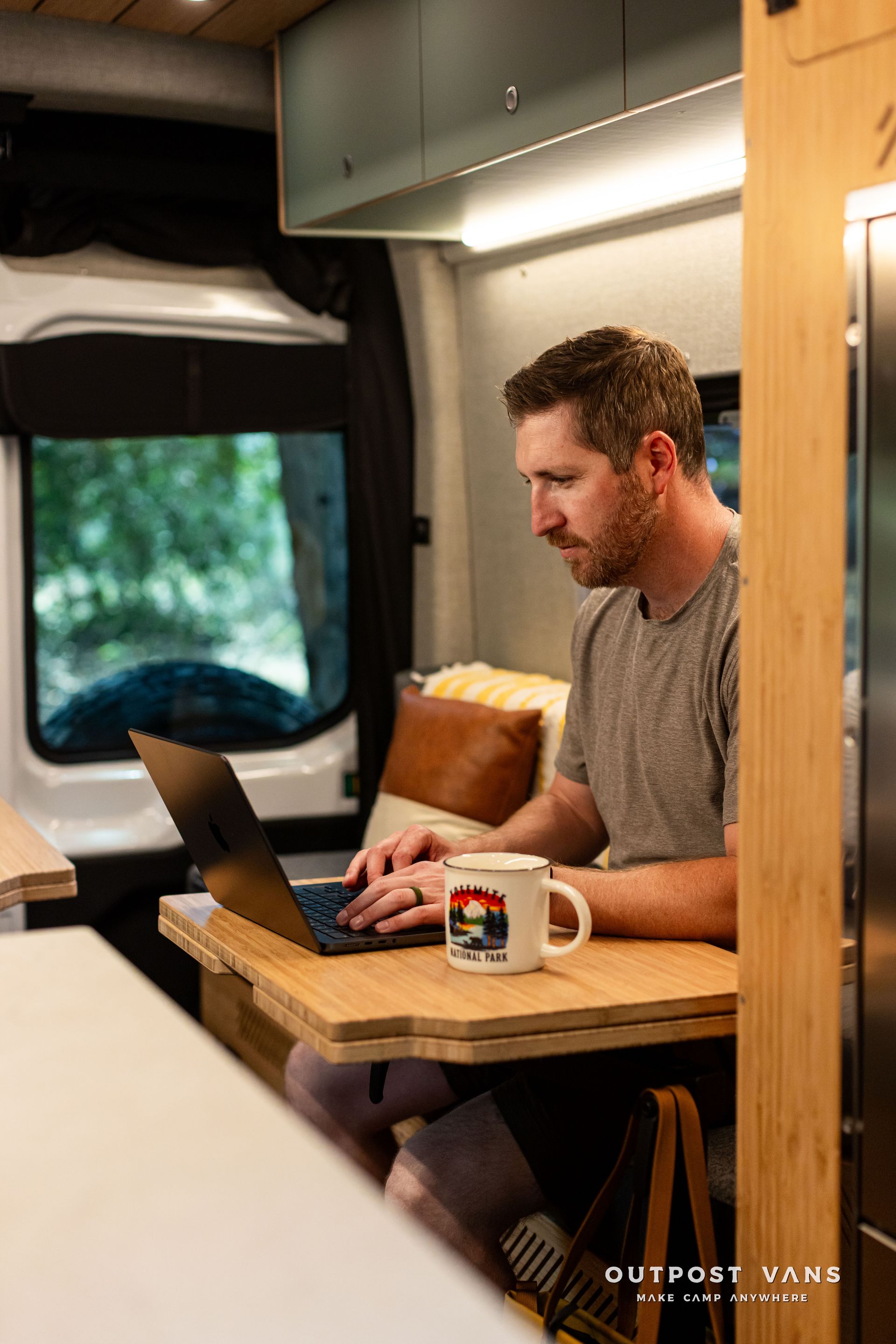 Man working on a laptop at a small wooden desk inside a camper van, with a mug nearby