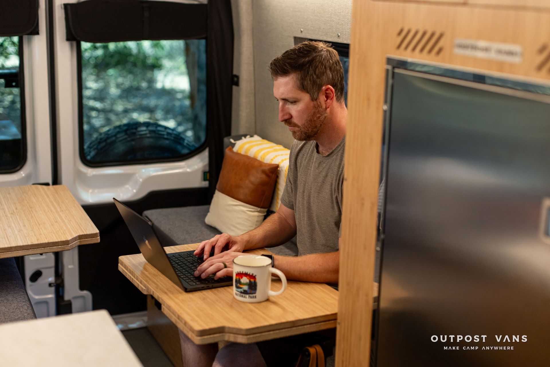 Man working on a laptop at a small table inside a van, with a mug beside him