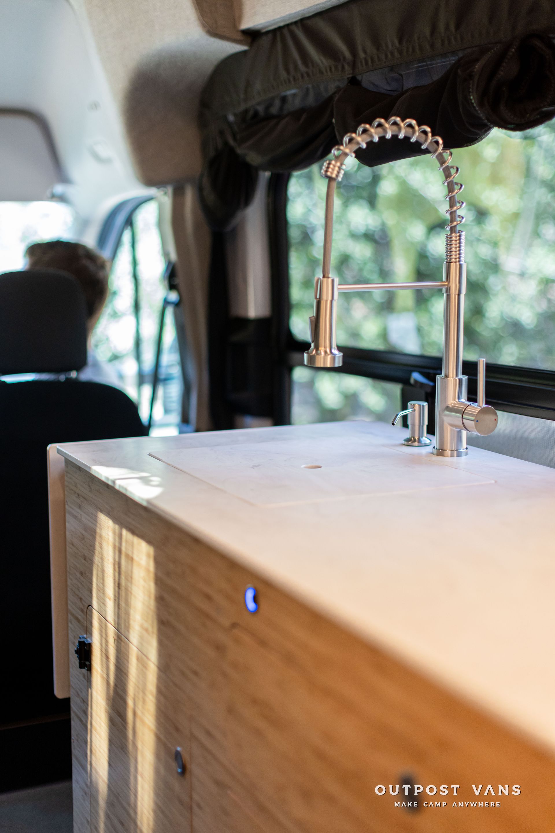 White countertop with faucet and sink inside a van, with a window showing greenery outside