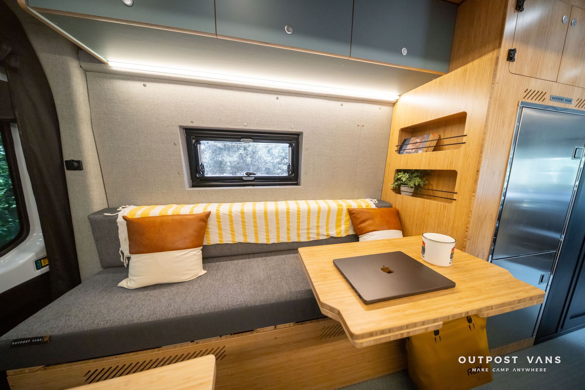 Modern camper van interior with a gray bench bed, orange cushions, and a laptop on a wooden table