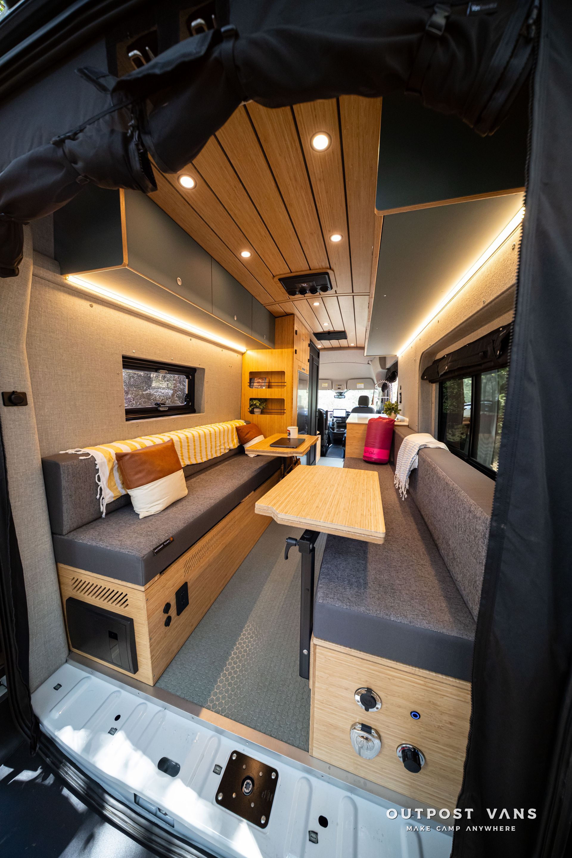 Narrow camper van interior with wood ceiling, gray seating, and a small table beside a bunk area.