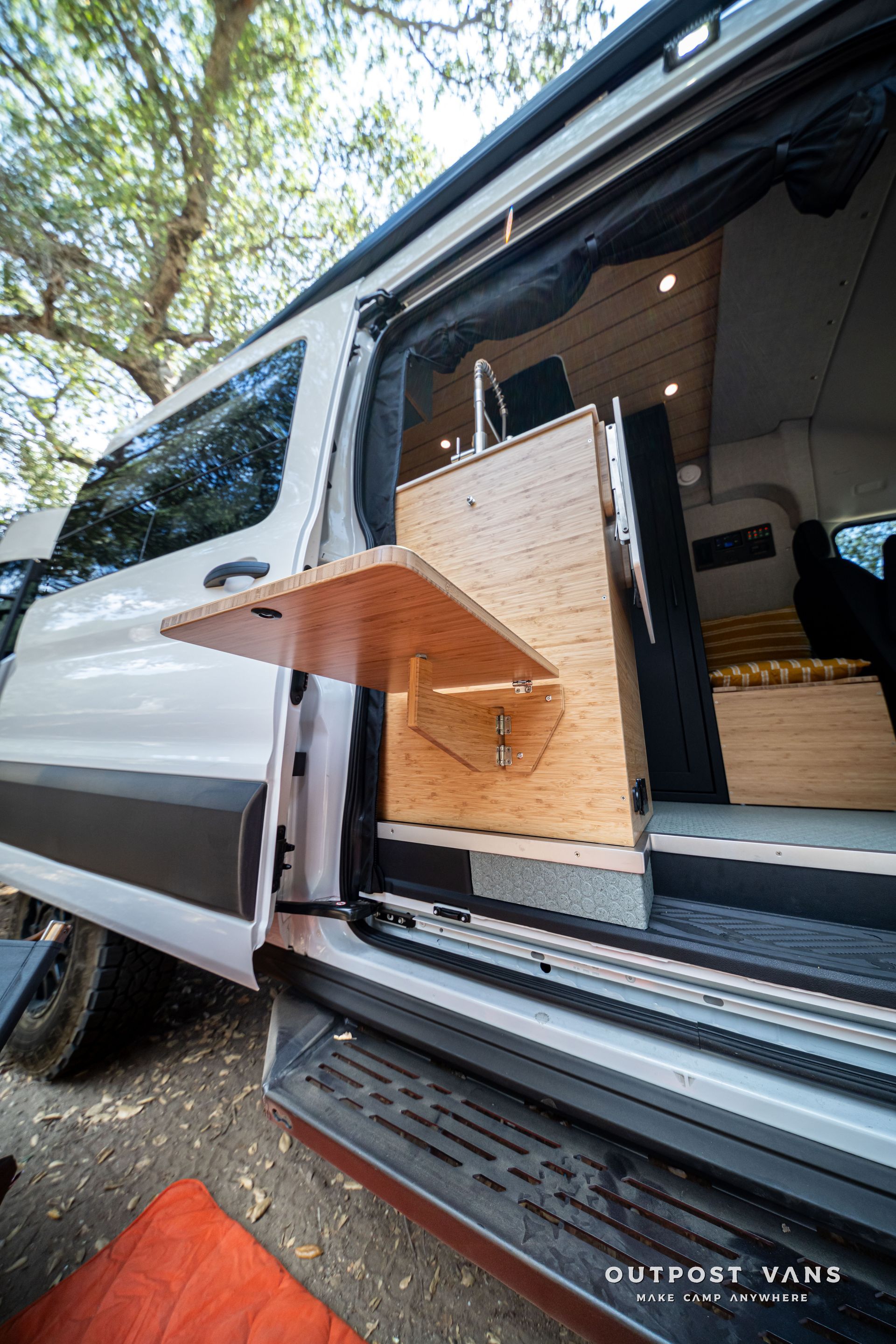 White camper van with a wooden sliding shelf extended from the side door in a wooded area