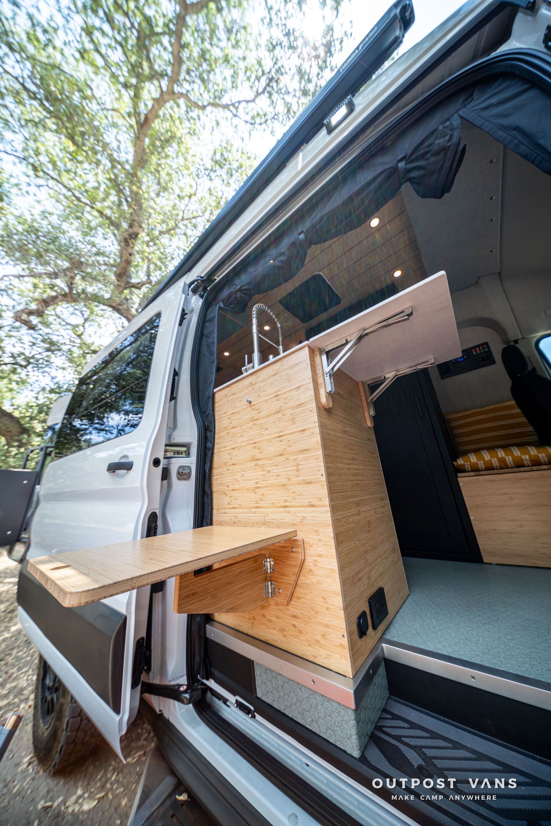 White camper van with open side door, showing a wooden interior setup and fold-out table under trees