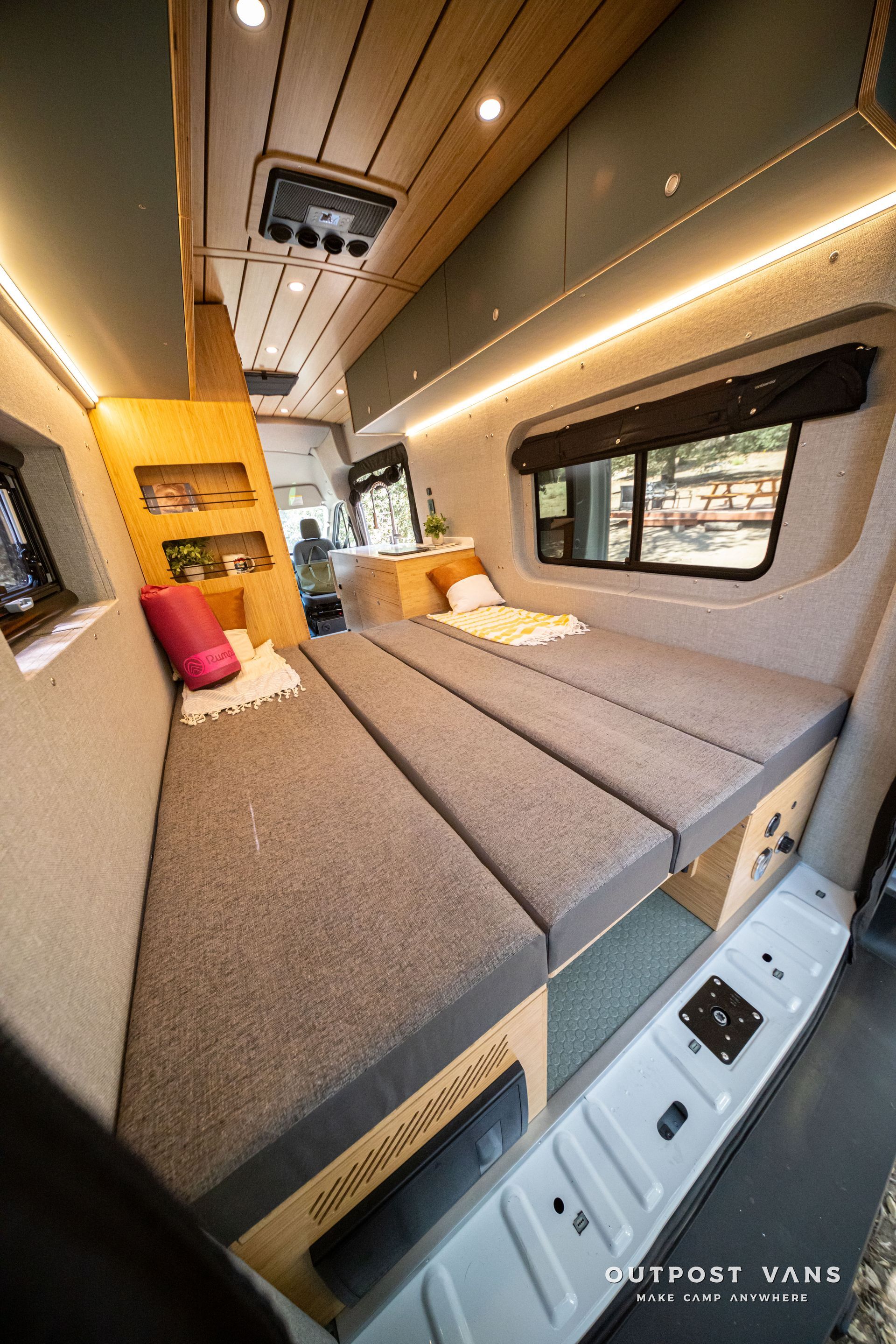 Compact camper van interior with gray bench bed, wood cabinets, skylight, and window seat