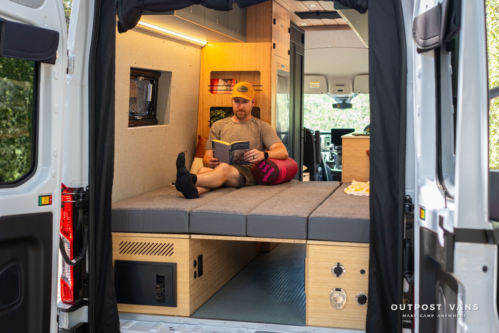 Man sitting on a bed in a compact camper van interior with warm lights and open rear doors