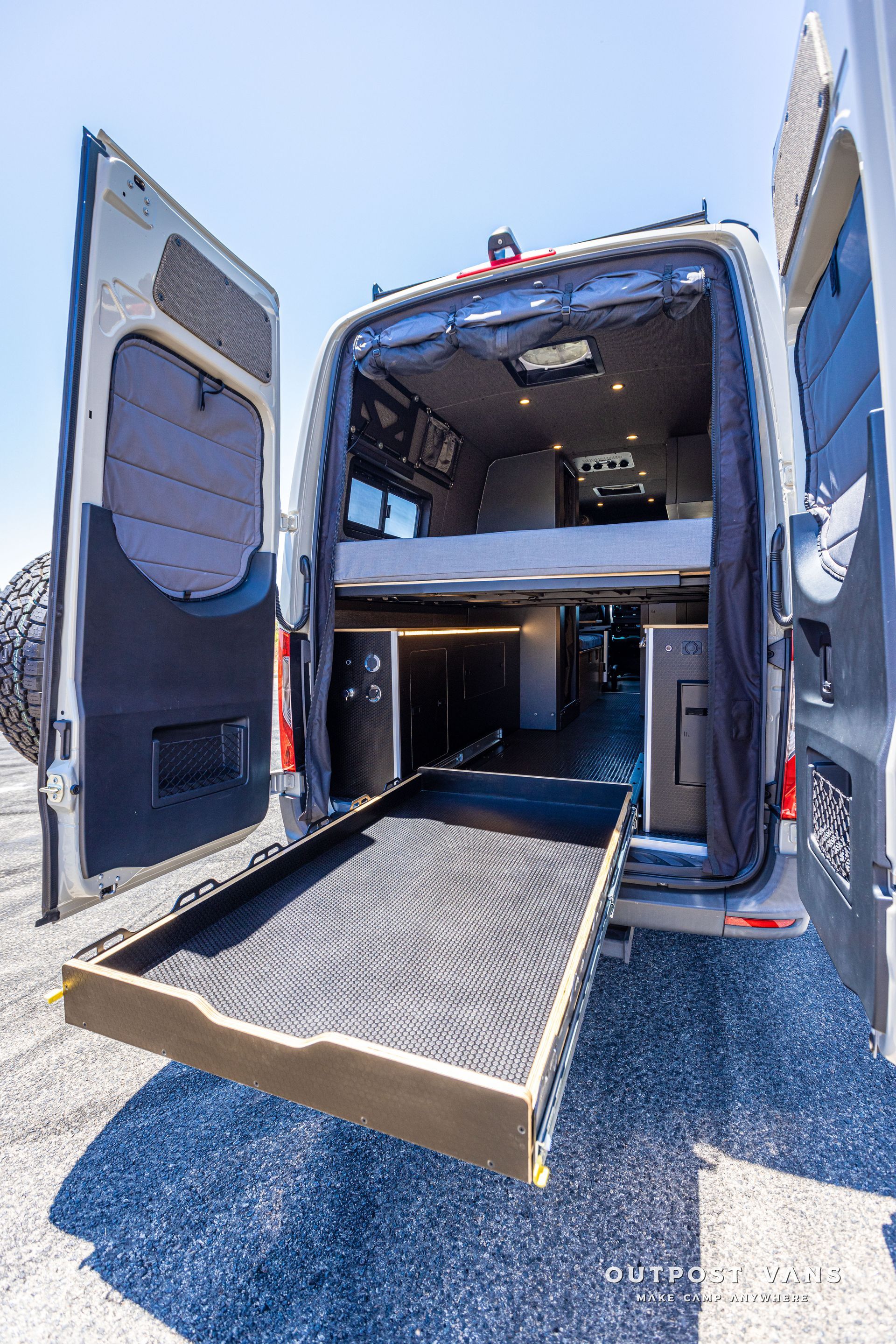 The back of a van with the door open and a bed in the back.