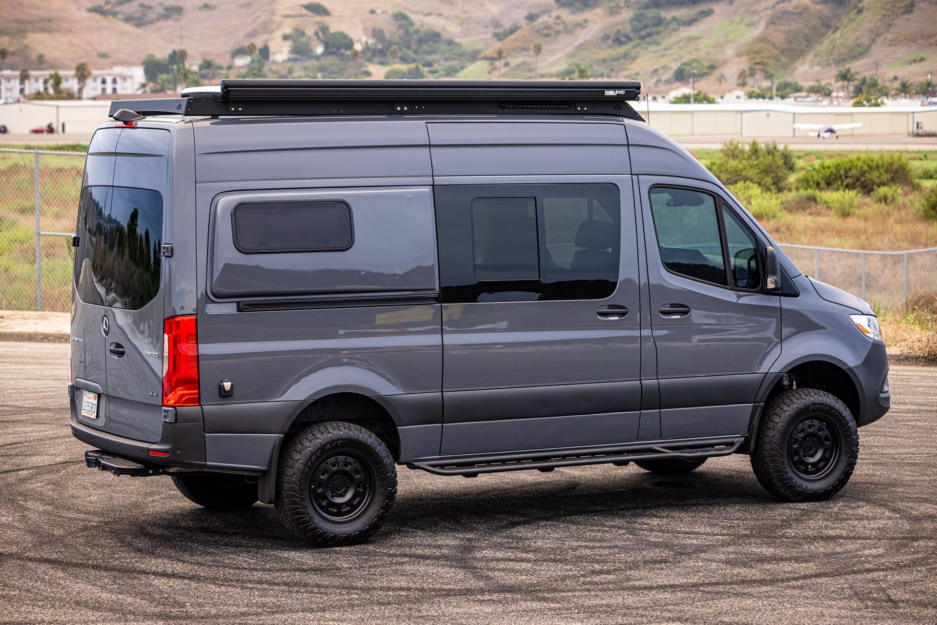 A gray van with a roof rack is parked on a dirt road.