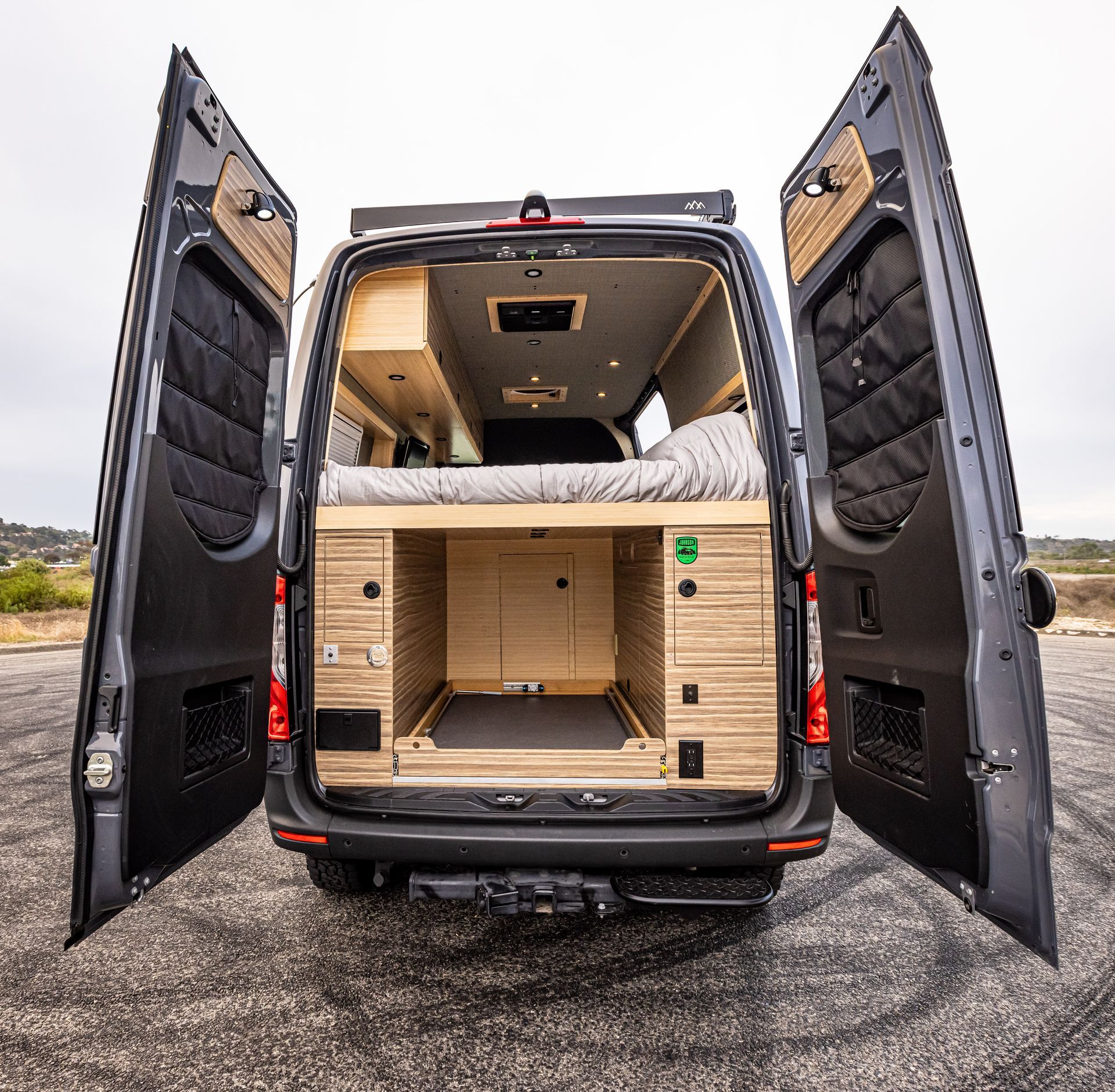 The back of a van with its doors open and a bed in the back.