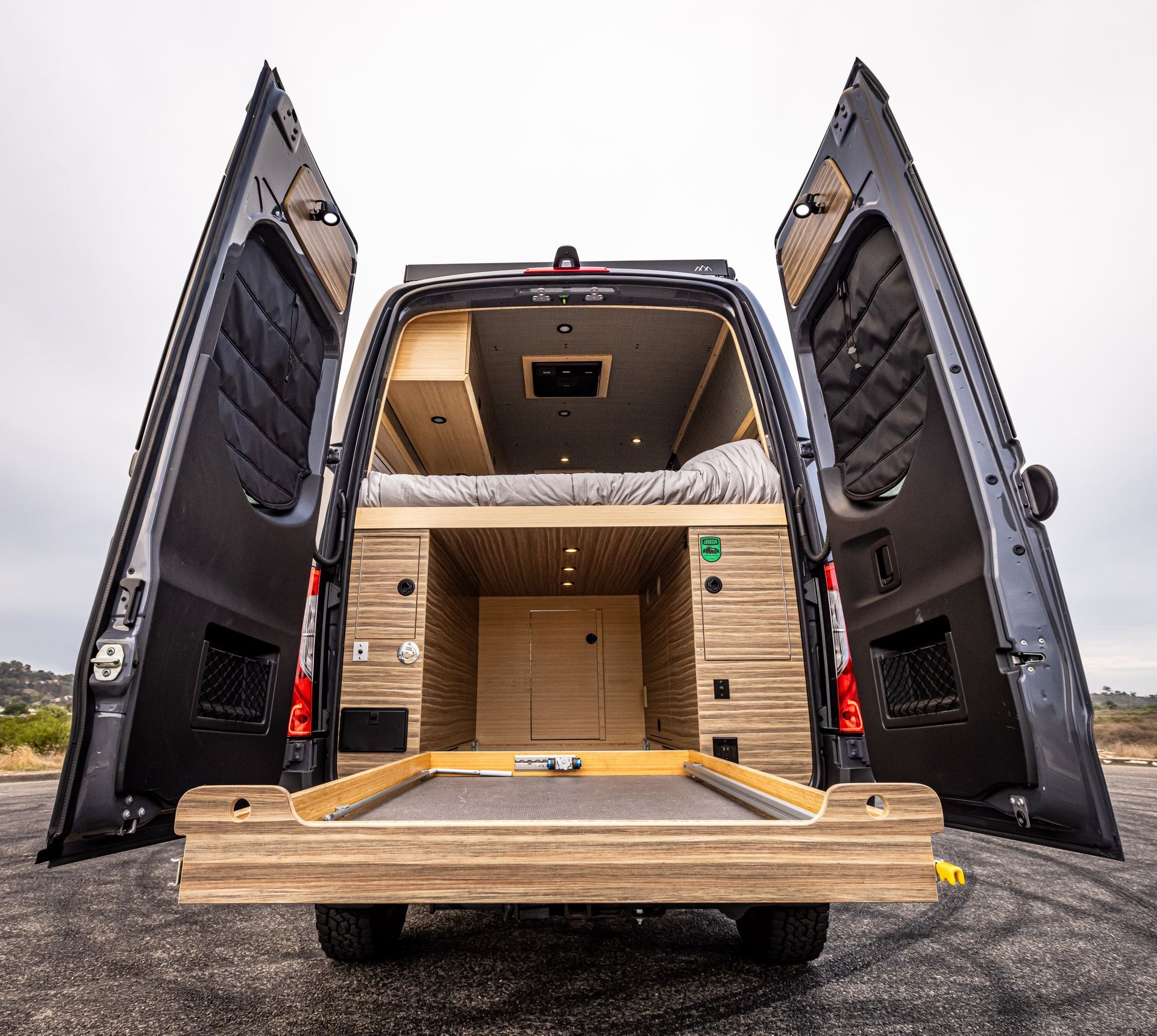 The back of a van with its doors open and a bed in the back.