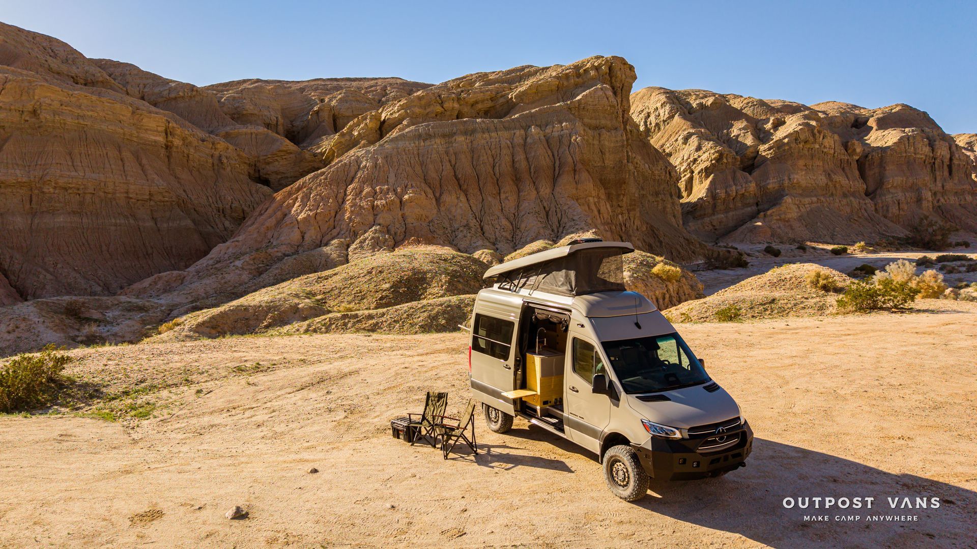 Camper van parked on sandy desert ground beside layered red rock cliffs under clear skies