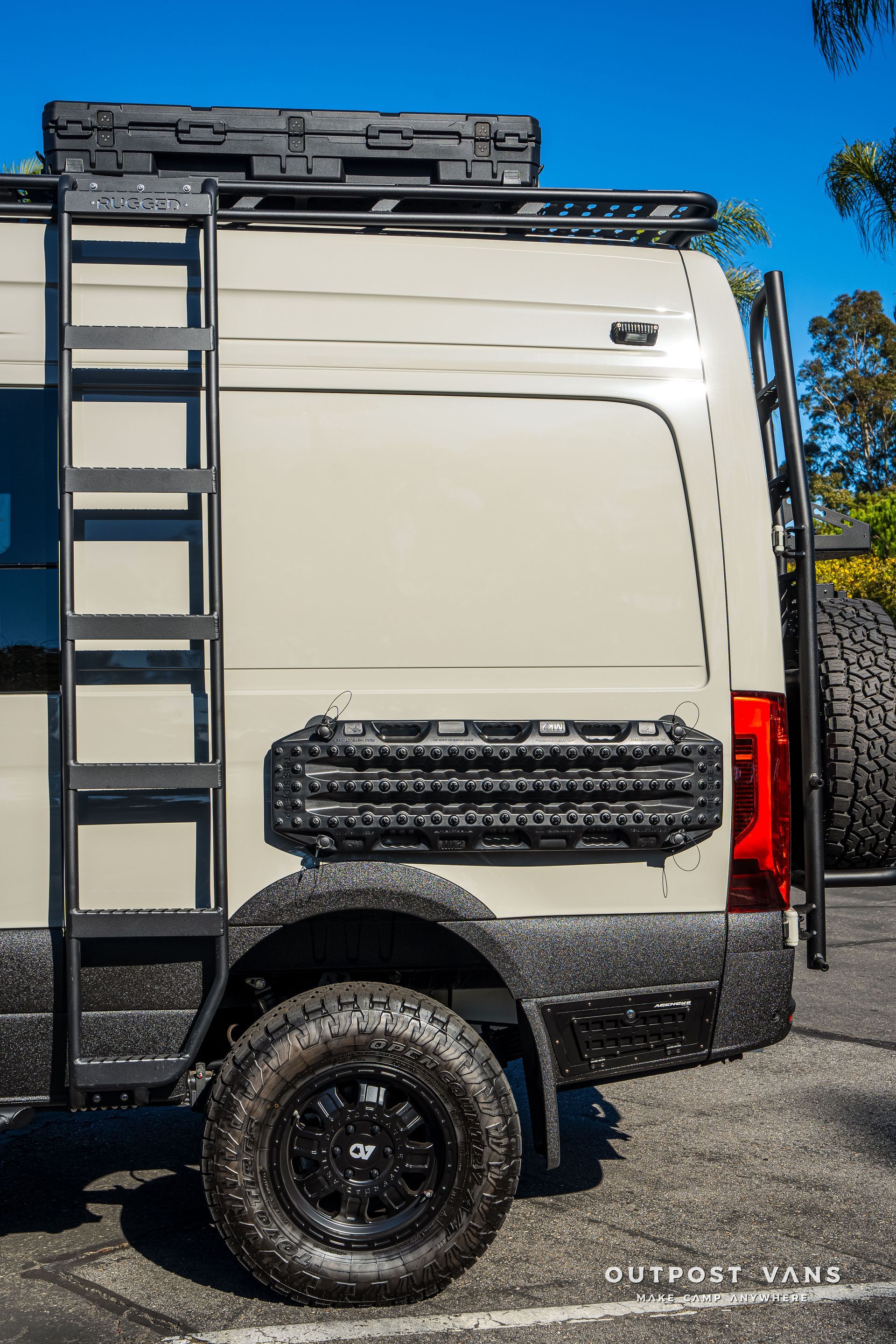 Tan camper van with black accessories: ladder, tire, storage cases, and rack.