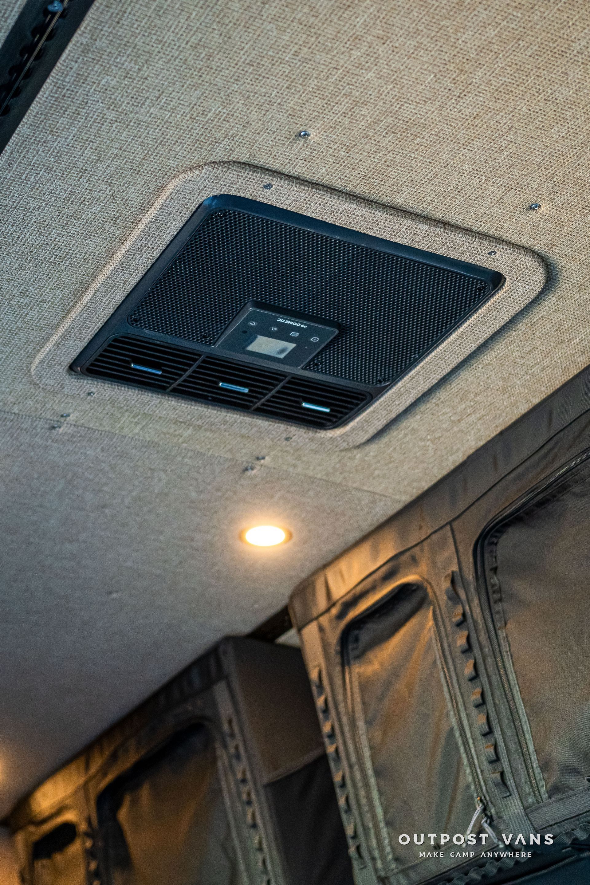 Overhead air vent with control panel in a vehicle, surrounded by textured fabric and recessed lighting.