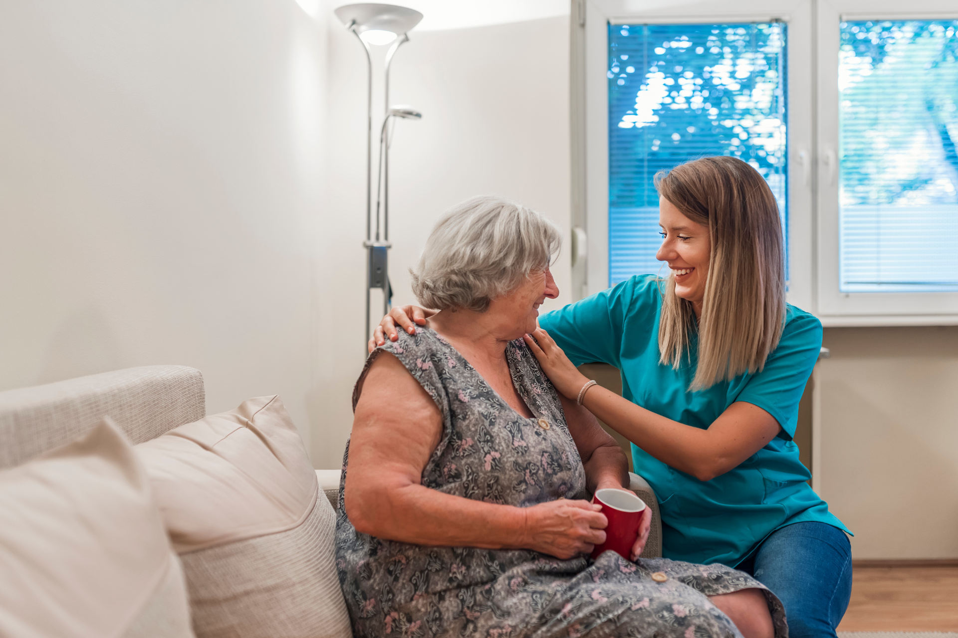 Caregiver consoling an elderly woman on a couch in a bright living room