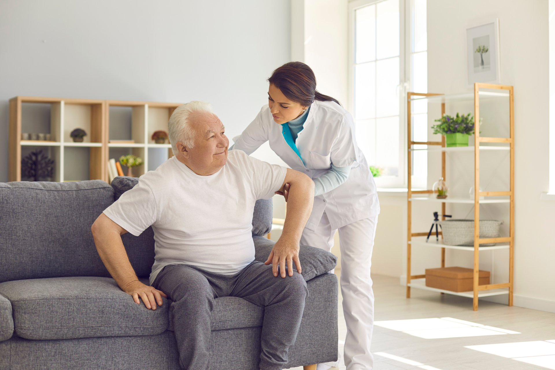 Caregiver helping an older adult stand from a gray sofa in a bright living room
