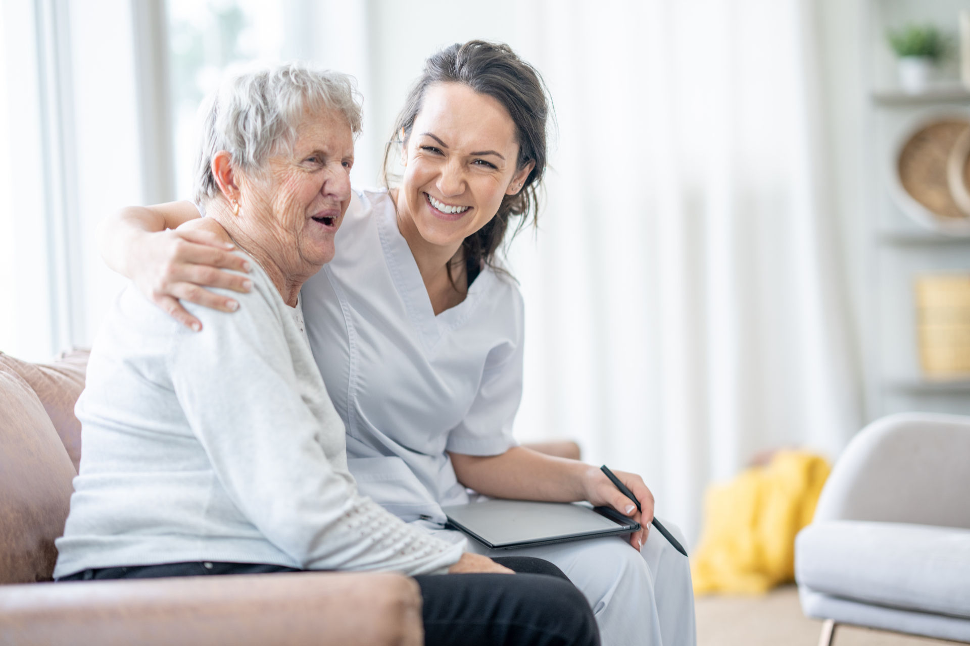 Smiling caregiver sitting with an elderly woman on a couch in a bright living room