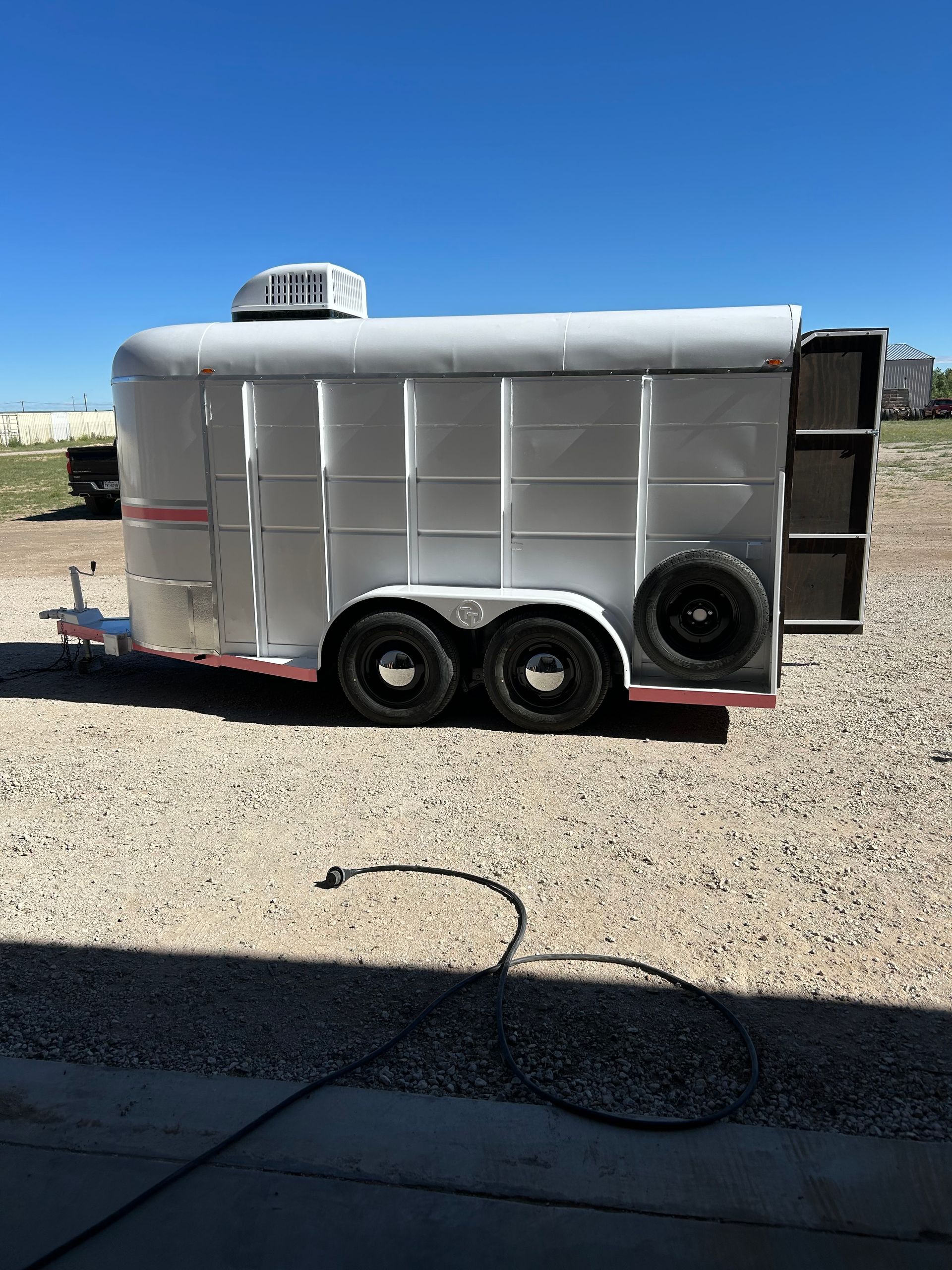 White enclosed trailer with dual axles and spare tire parked on gravel under a clear blue sky