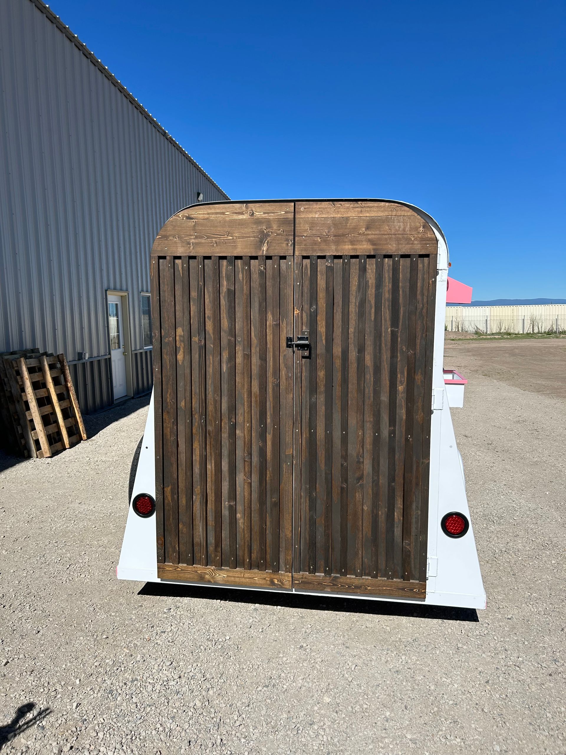 Small enclosed trailer with a weathered wooden rear door parked on gravel beside a metal building.