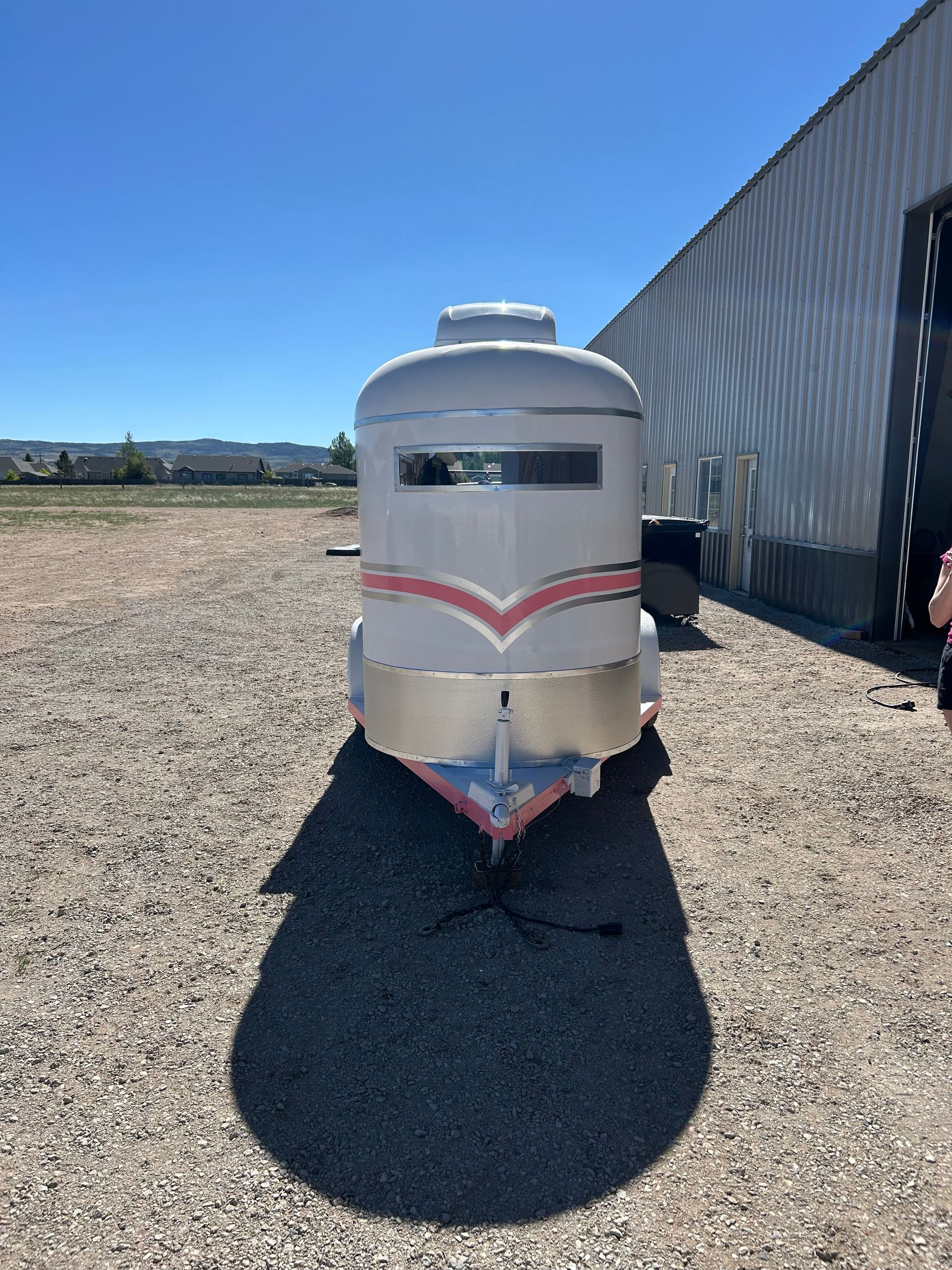 White horse trailer with red stripe parked on gravel beside a metal building under a clear blue sky