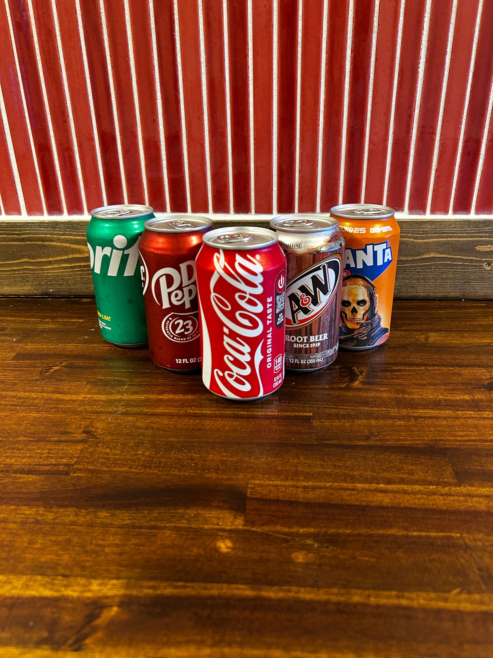 Five colorful soda cans lined up on a wooden shelf against a warm pink-lit background