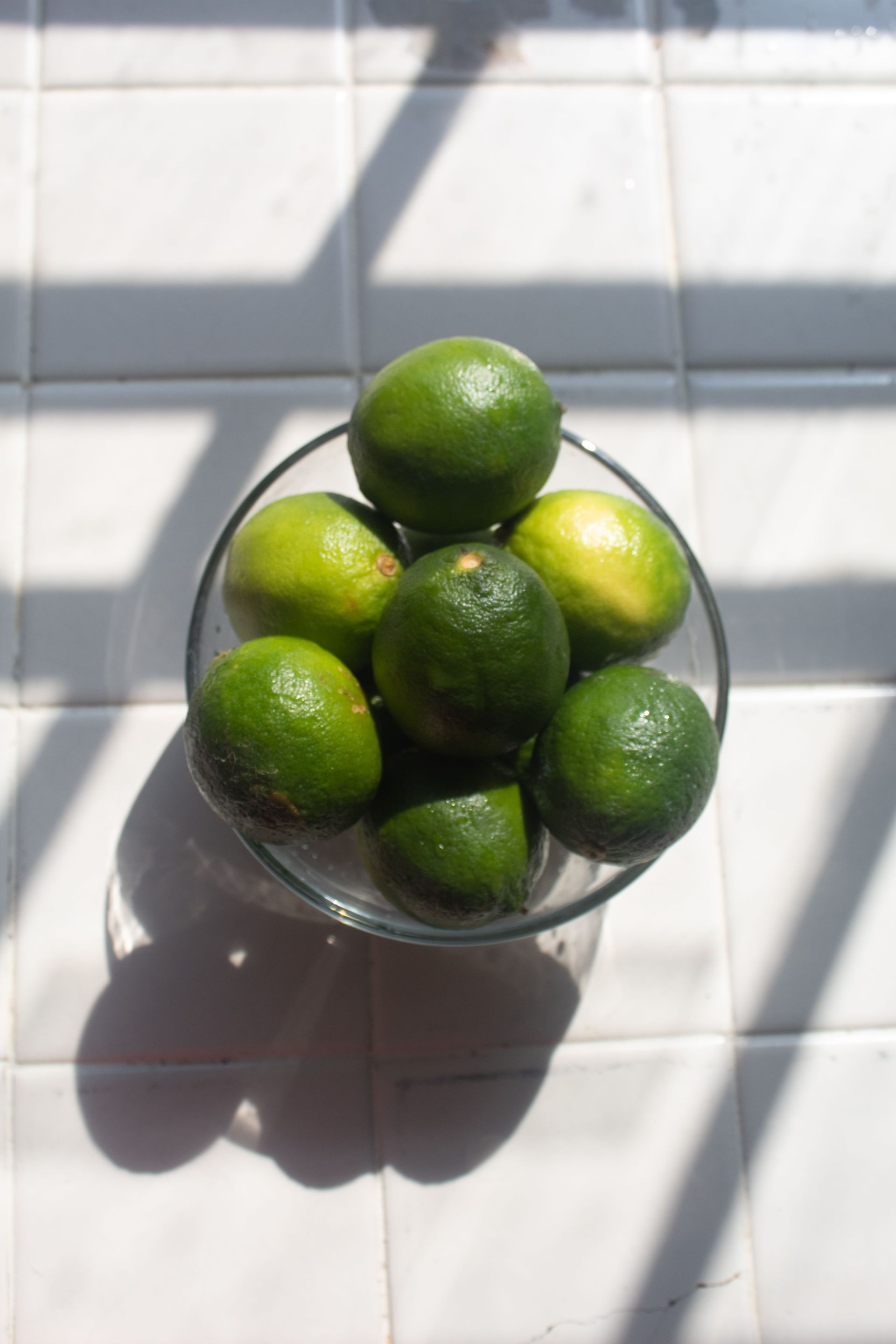 Bowl of green limes on a sunlit tiled surface, casting a shadow