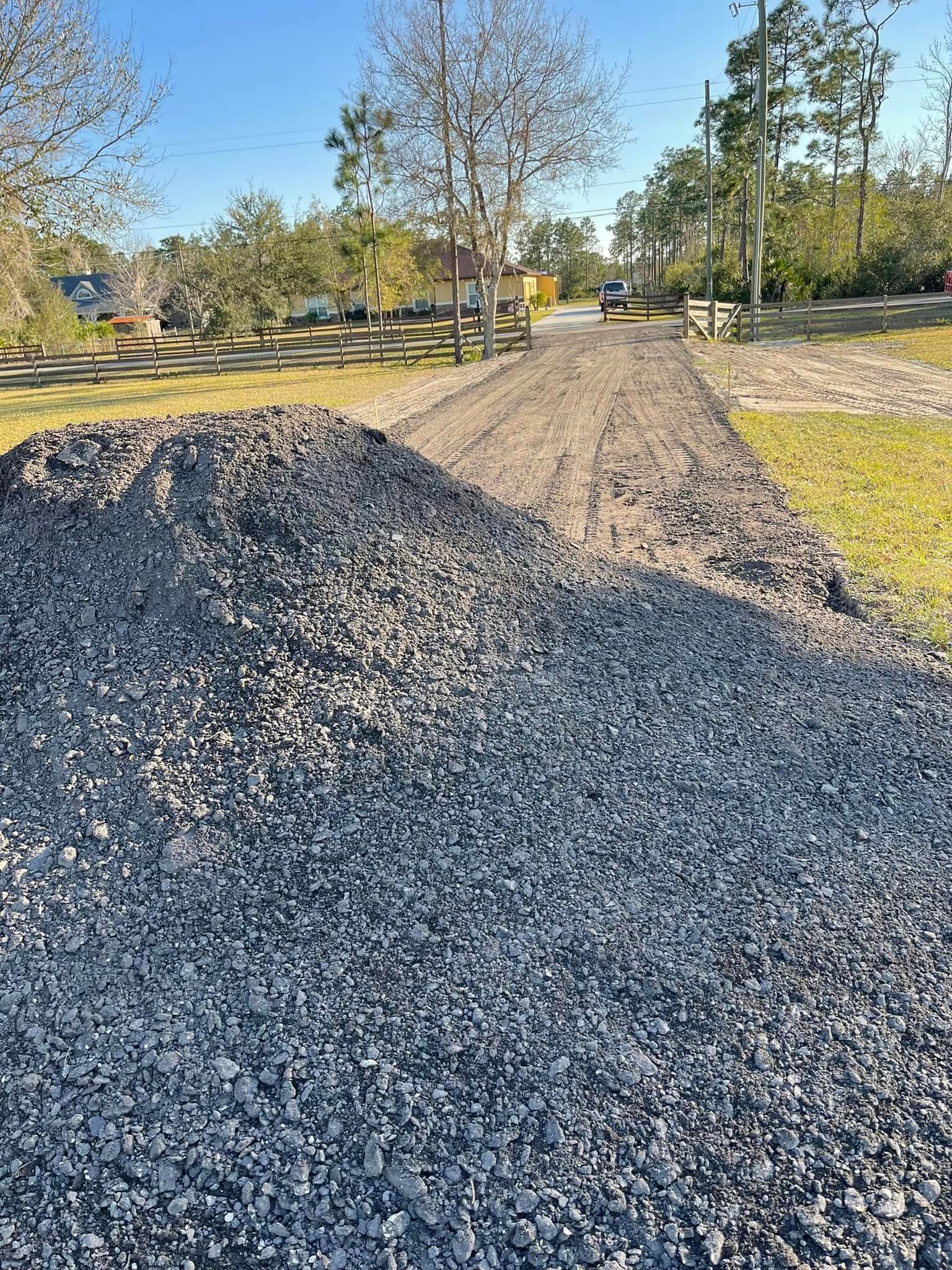Gravel piles beside a gravel driveway under a clear blue sky, with trees and grass in the background