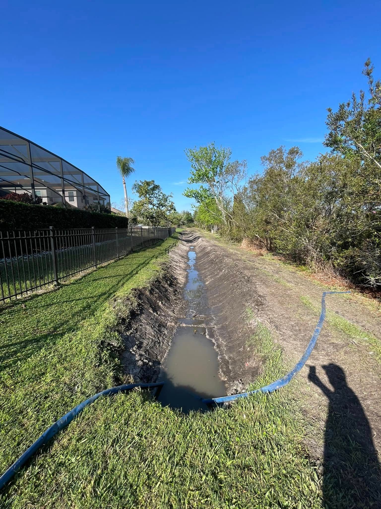 Narrow muddy drainage canal beside grass and shrubs under a clear blue sky, with a person’s shadow in the foreground.