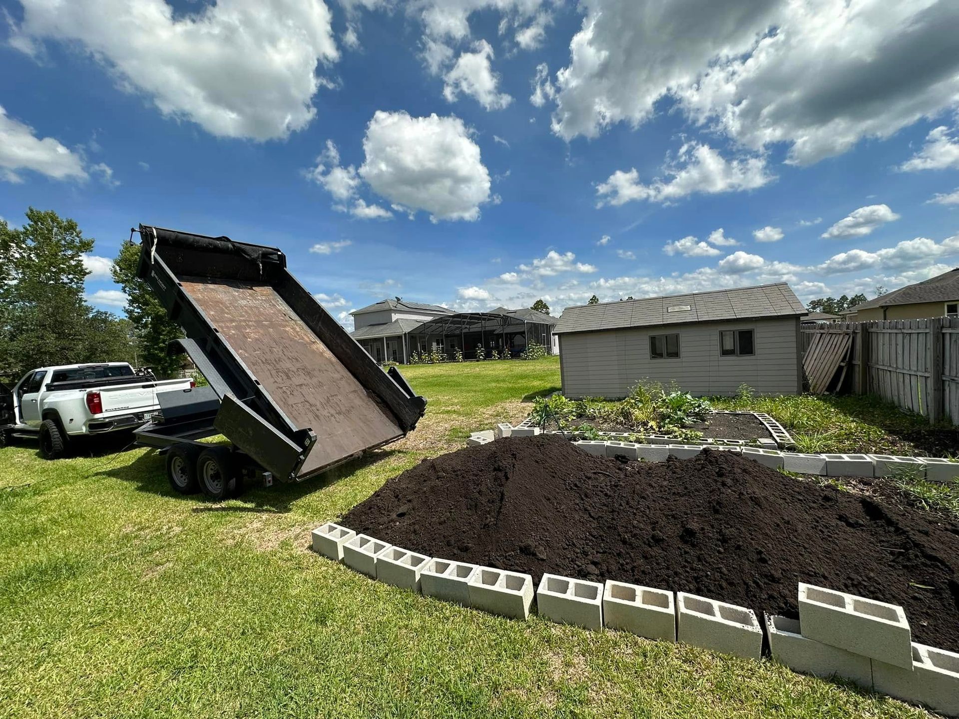 Dump truck unloading dark soil beside a fenced garden in a sunny backyard.