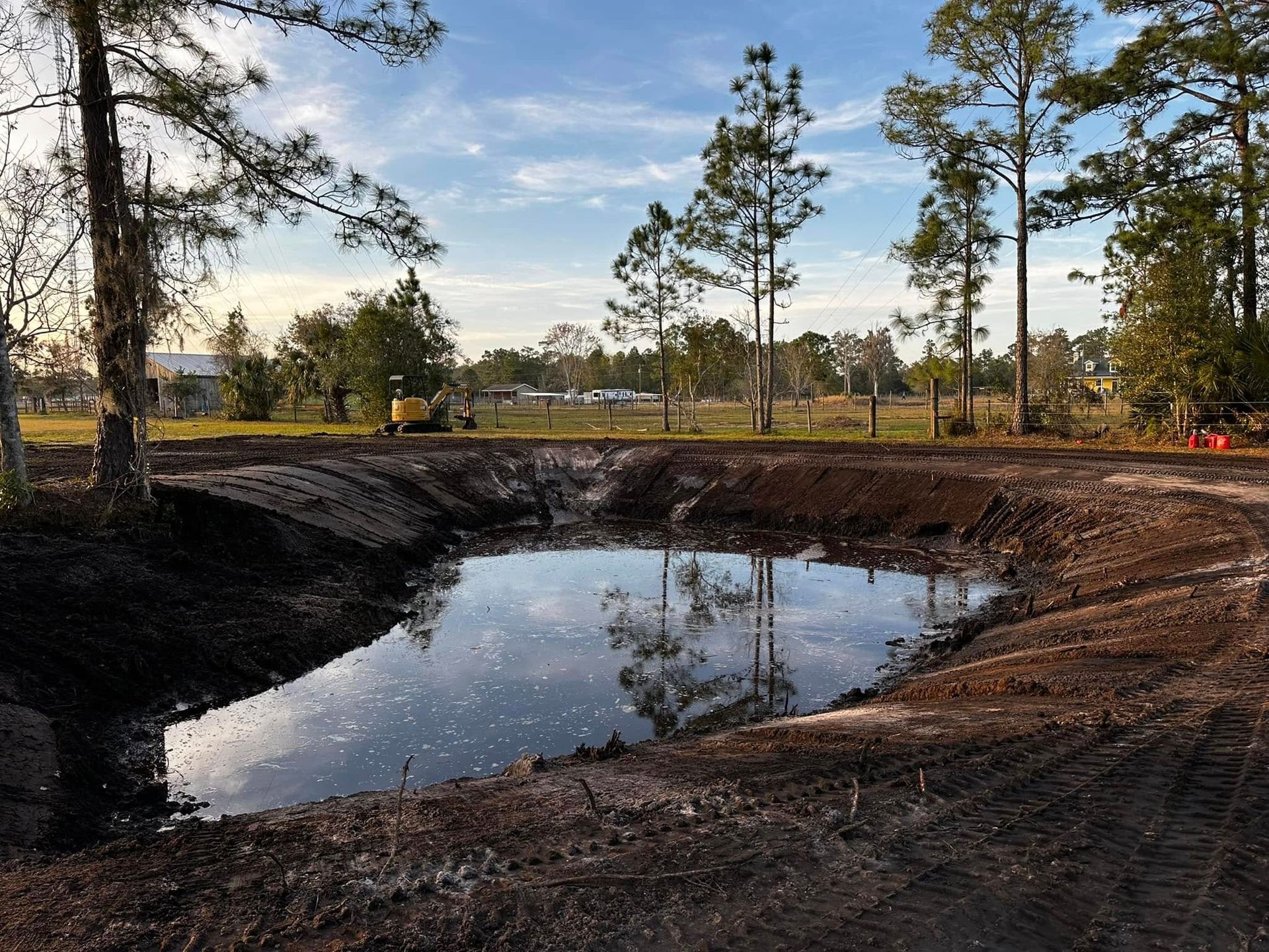 A muddy excavation pond reflecting pine trees under a clear sky at a construction site