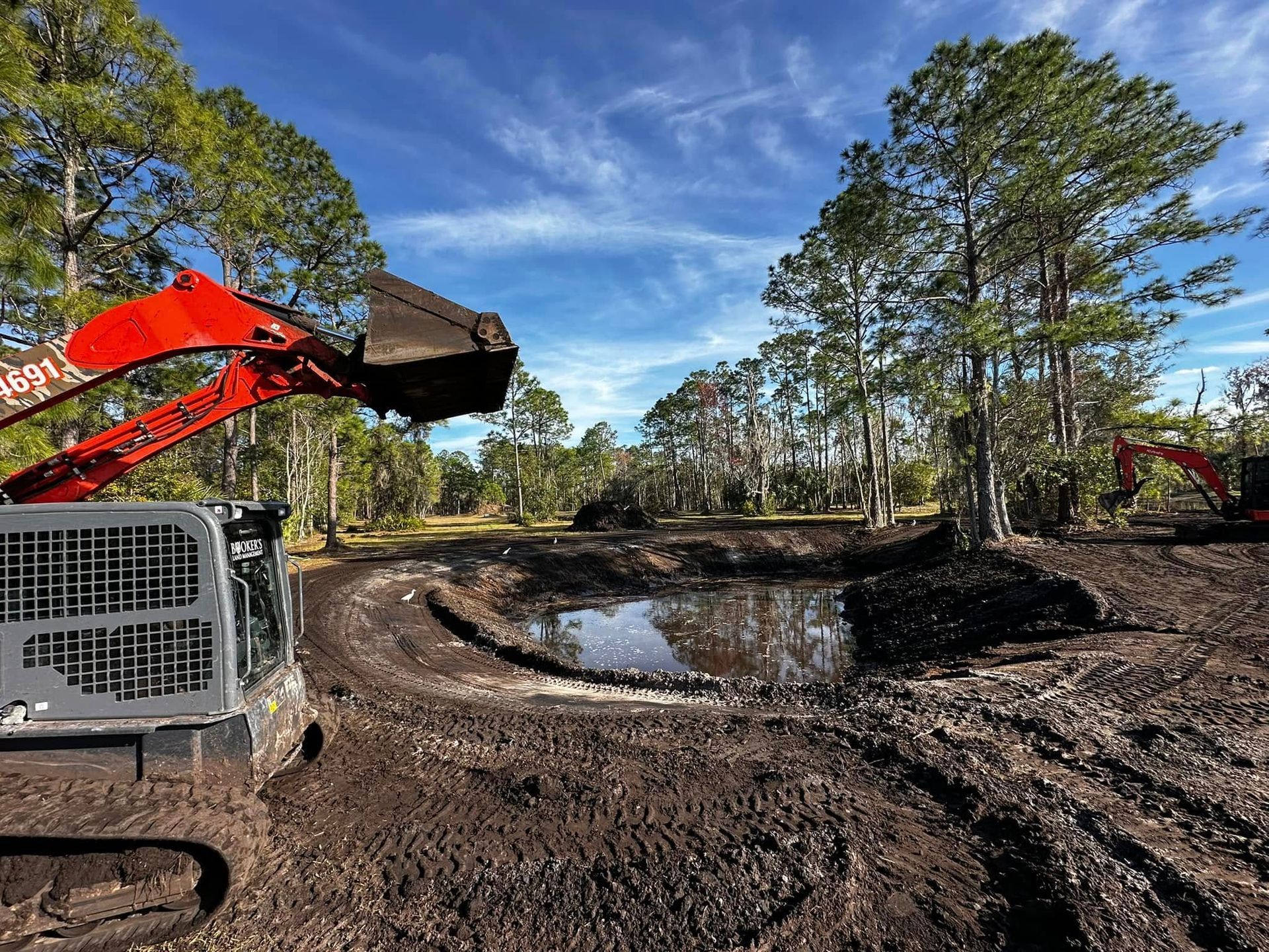 Excavator working on a muddy construction site with puddles, trees, and a blue sky.