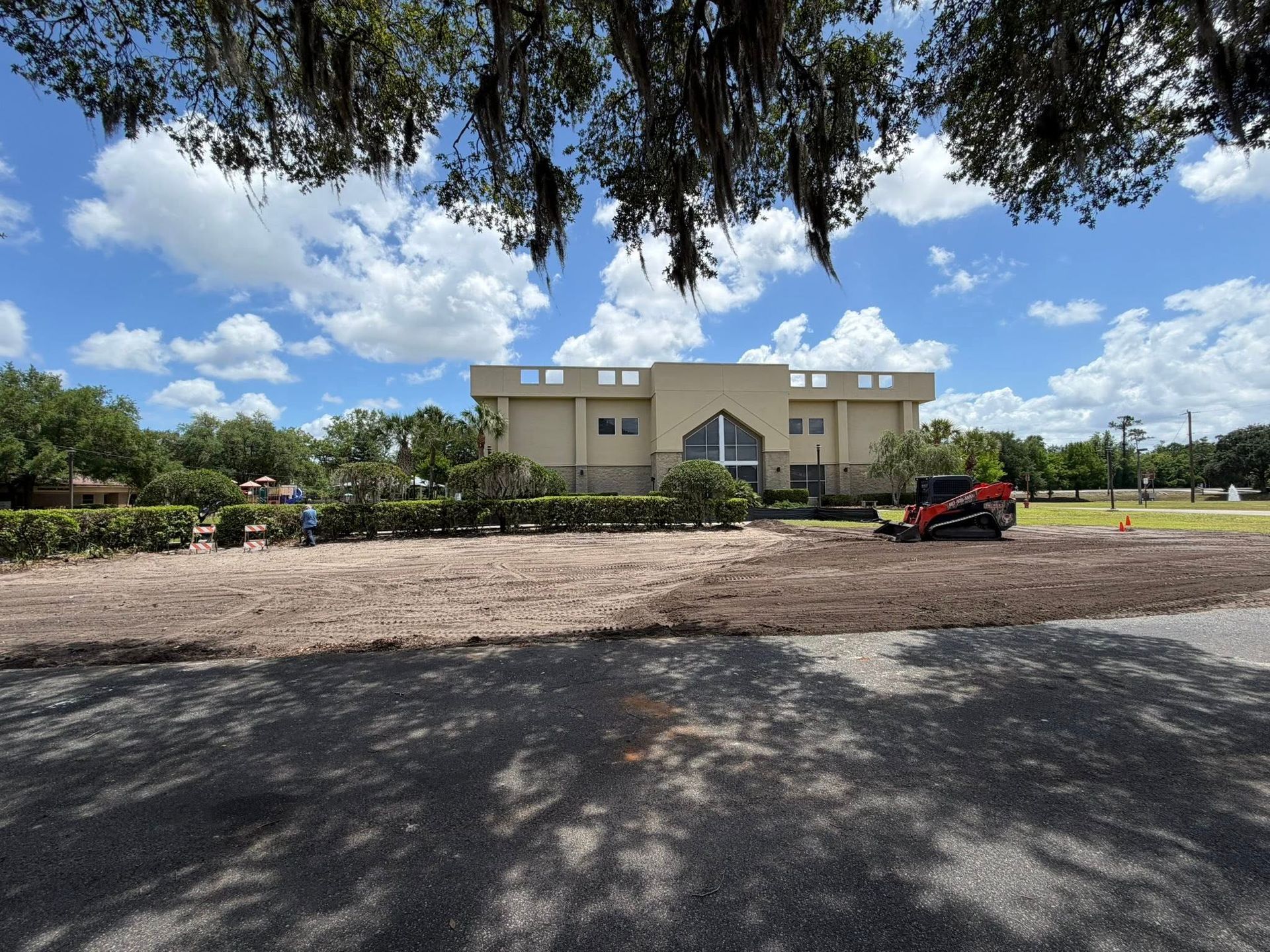 Beige building behind a gravel lot, with trees overhead and a red utility cart on the right.