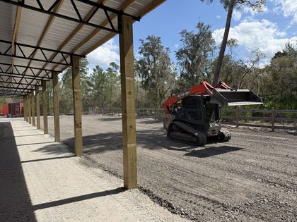 Covered outdoor work area with a small red-and-black tracked loader on gravel under a metal roof.